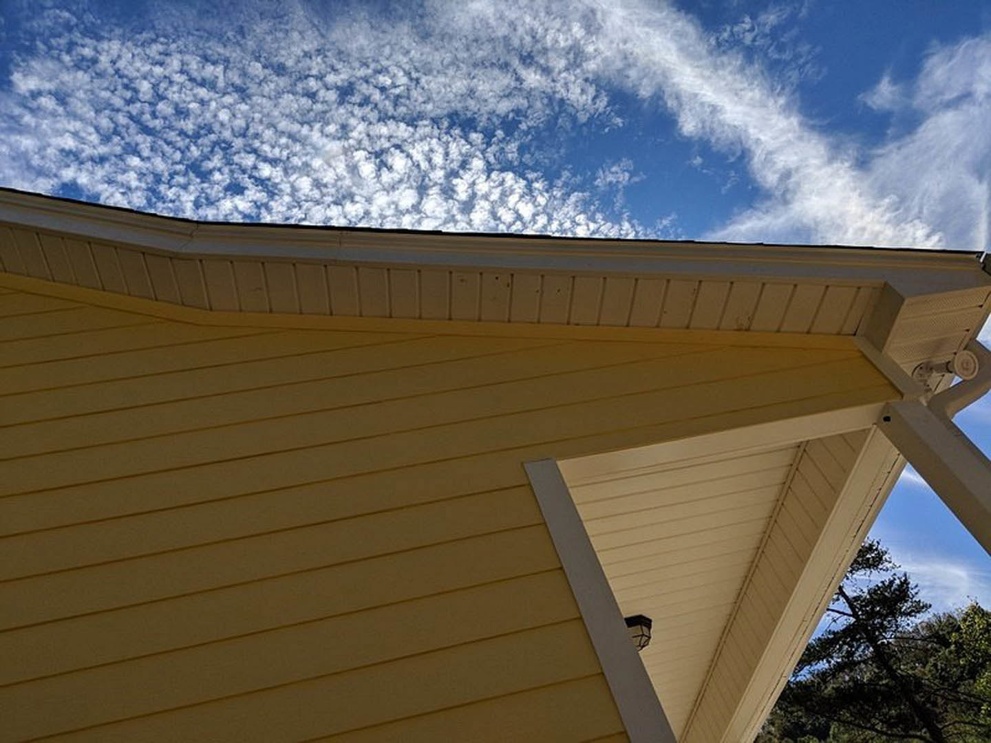 Yellow house with white roof under blue sky, surrounded by trees, featuring a black and white lamp and white wall with black stripe