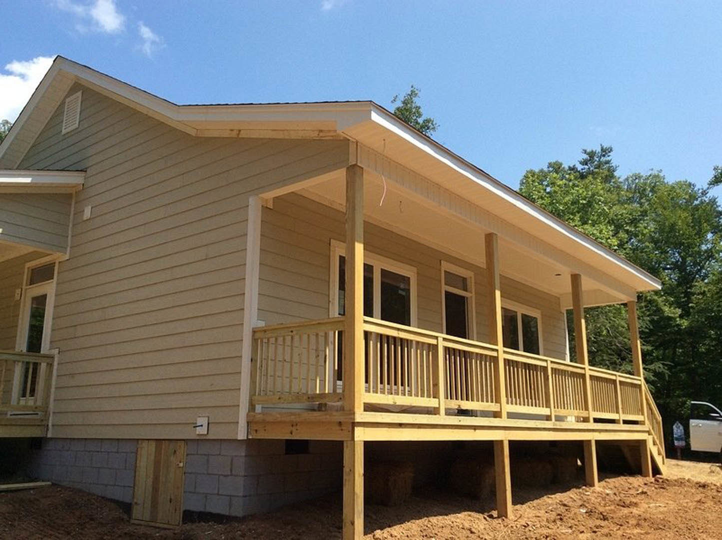 White siding home with covered wooden porch, railing, and deck; large windows, wooden front door, tile accent wall, and trees in the background.
