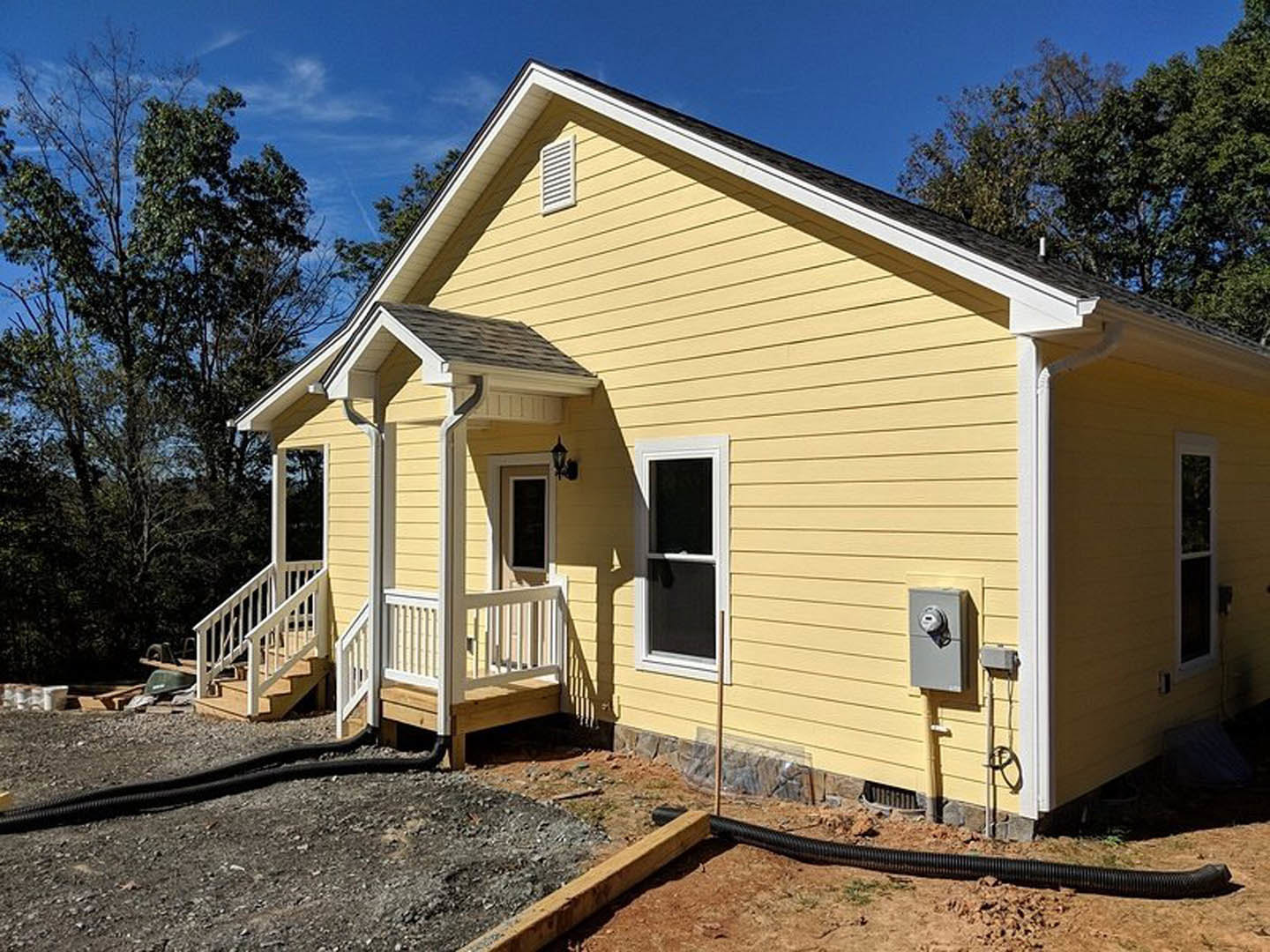 Yellow siding house with white porch railing, black hose coiled near entry, large windows, and staircase visible; leafy tree partially frames exterior.