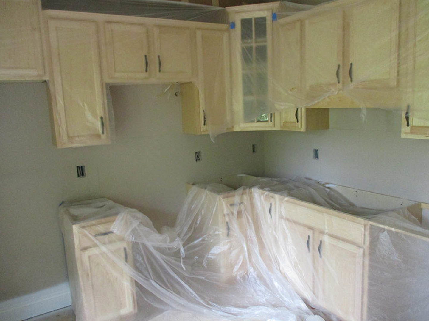 Kitchen with cabinets, countertops, and appliances covered in clear plastic wrap, white walls, and a blurry window in the background