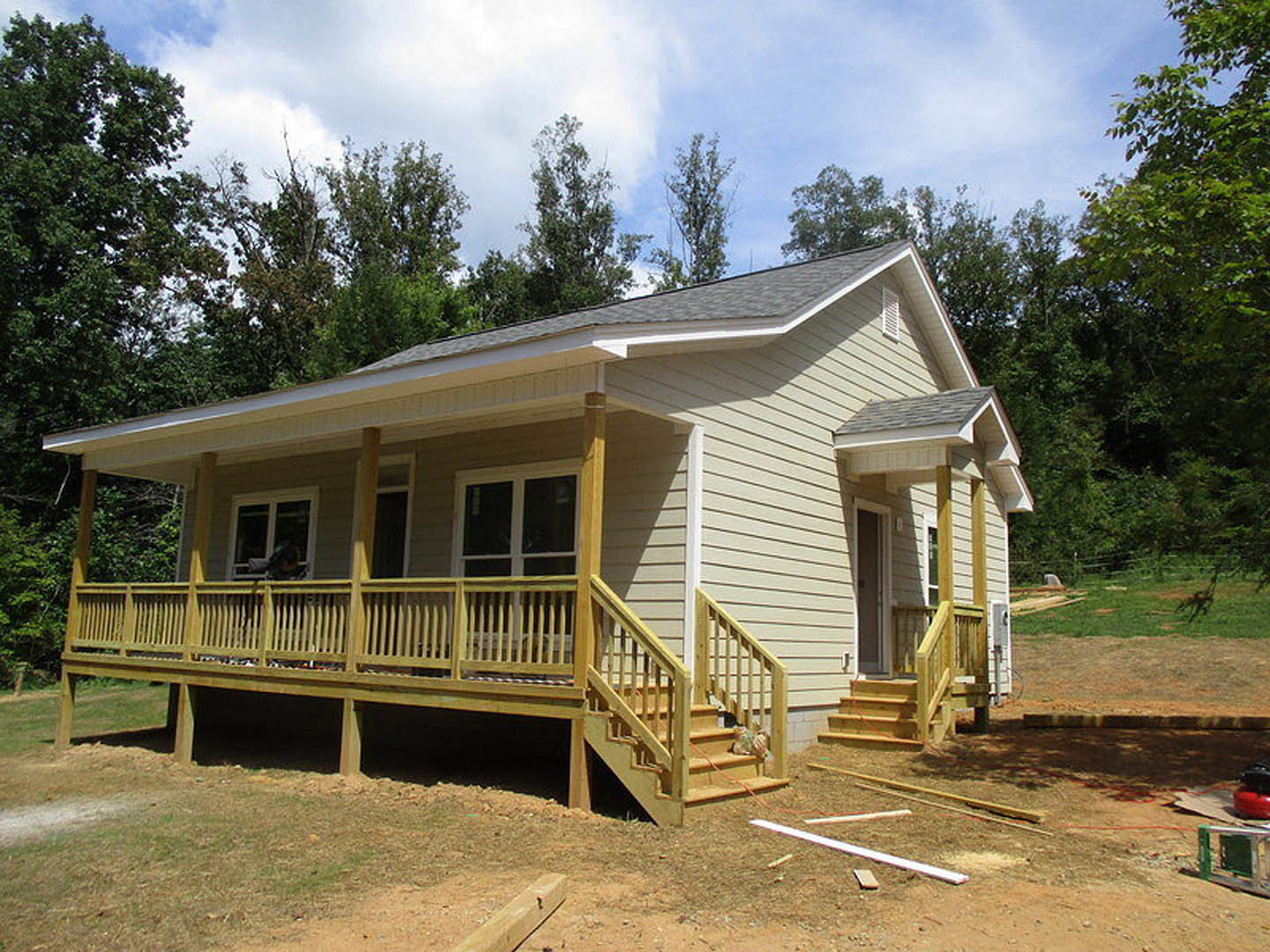 Partially built house with exposed wooden porch, unfinished siding, railing, and visible stairs, set against cloudy sky and trees.