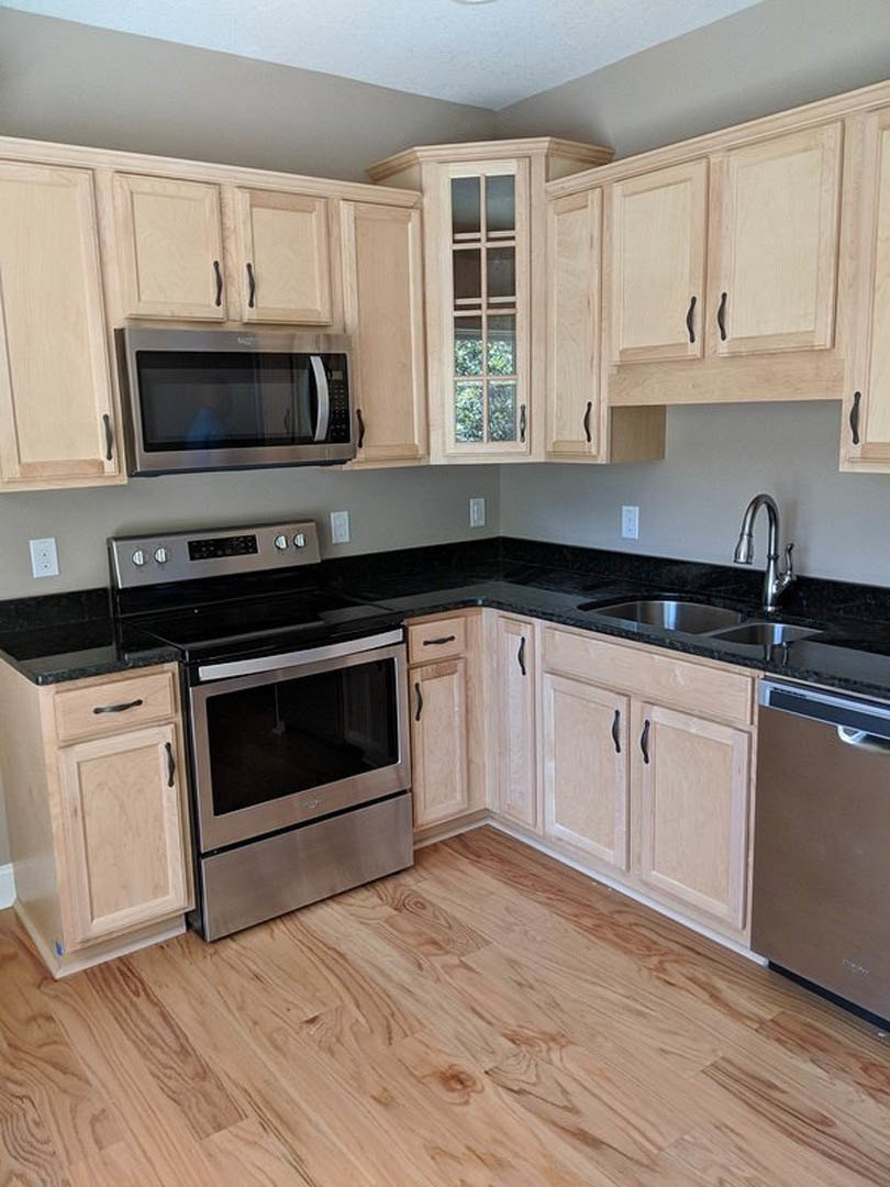 Wooden kitchen cabinets with matte black countertops, stainless steel oven, microwave, modern faucet, large window, and neutral wall finishes; person standing near microwave.