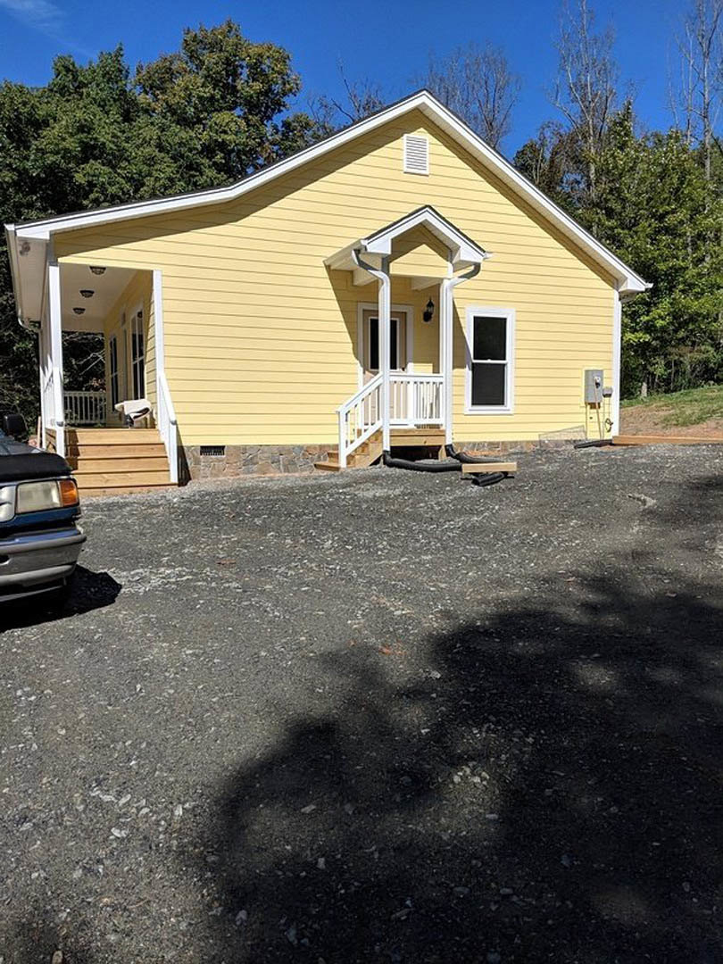 Yellow siding house with white trim and porch, parked car in driveway, tree shadows cast on lawn, screened window visible.