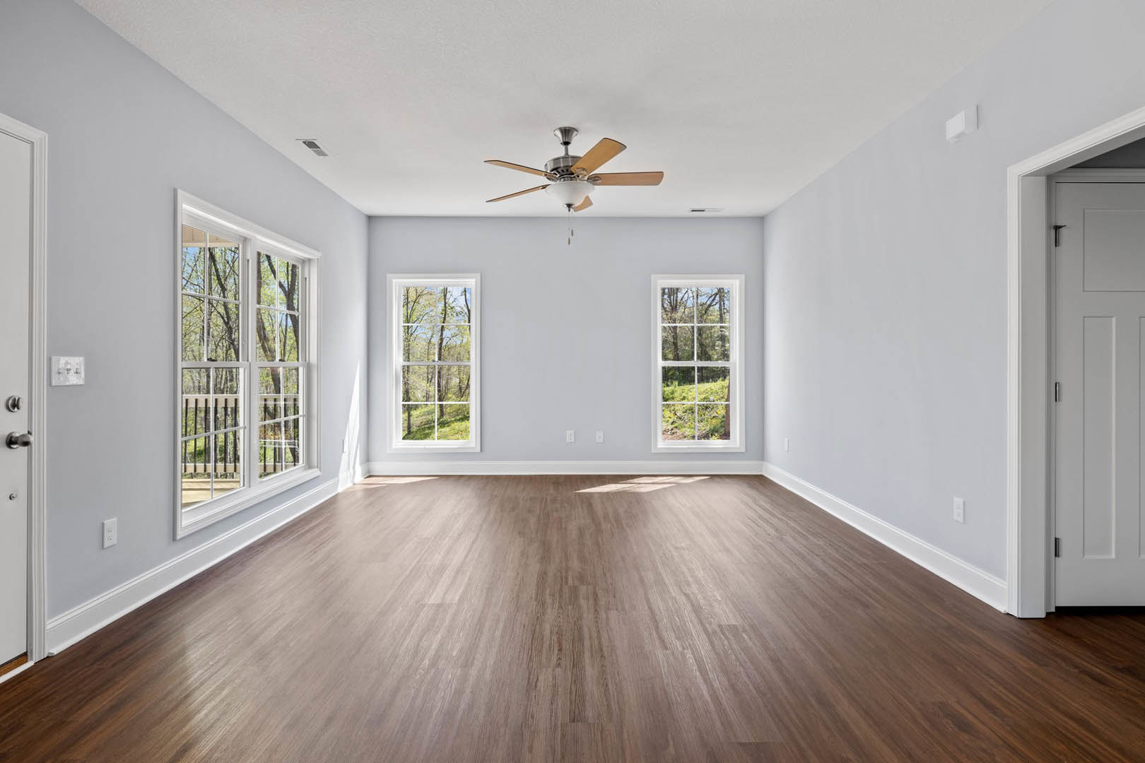 Wood flooring in a room with white walls, ceiling fan with light fixture, large windows showing trees and forest views, three switches on the wall
