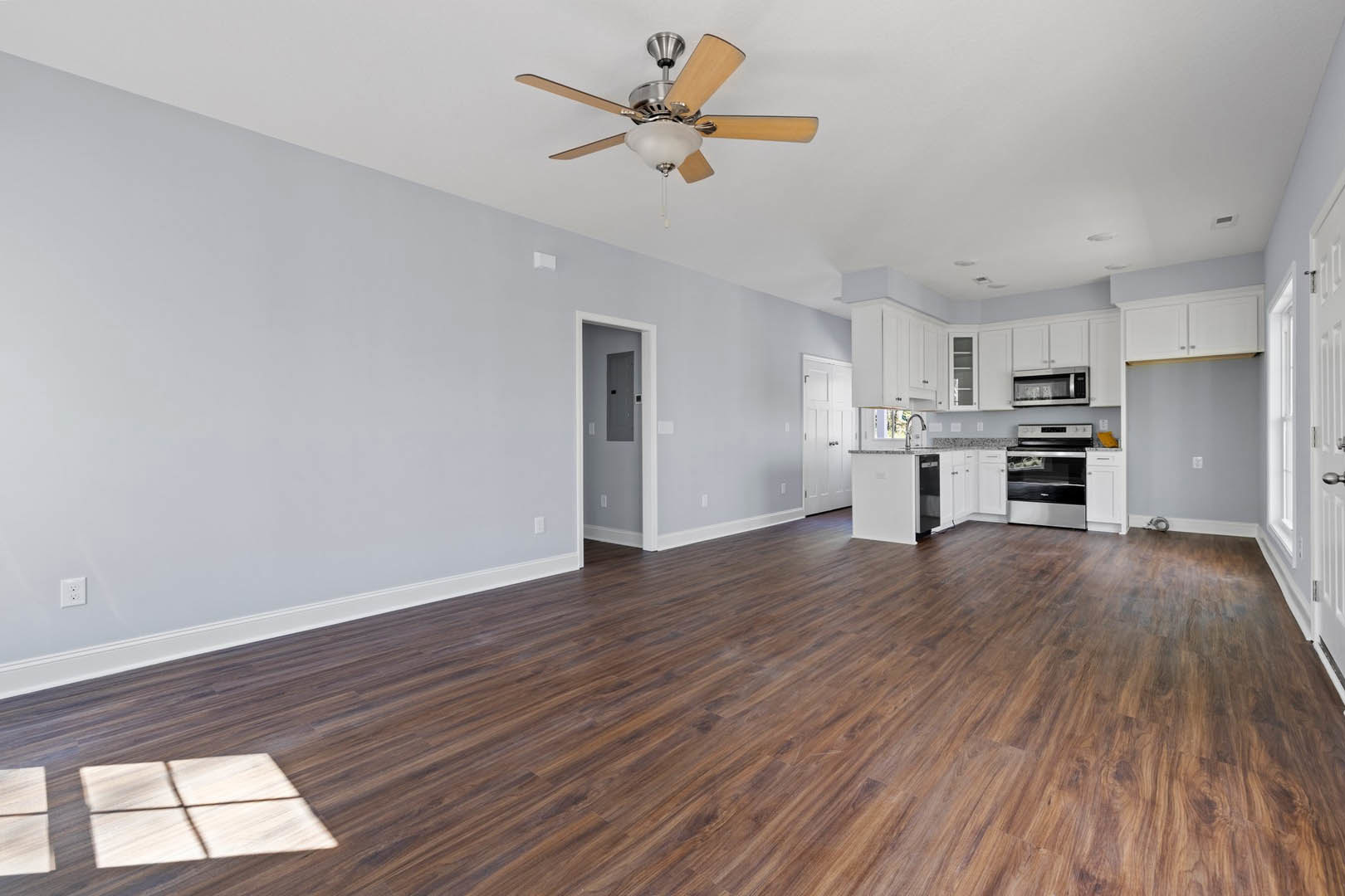 Open living space with hardwood flooring, white walls, ceiling fan with light fixture, stainless steel oven and microwave, white door with inset panel