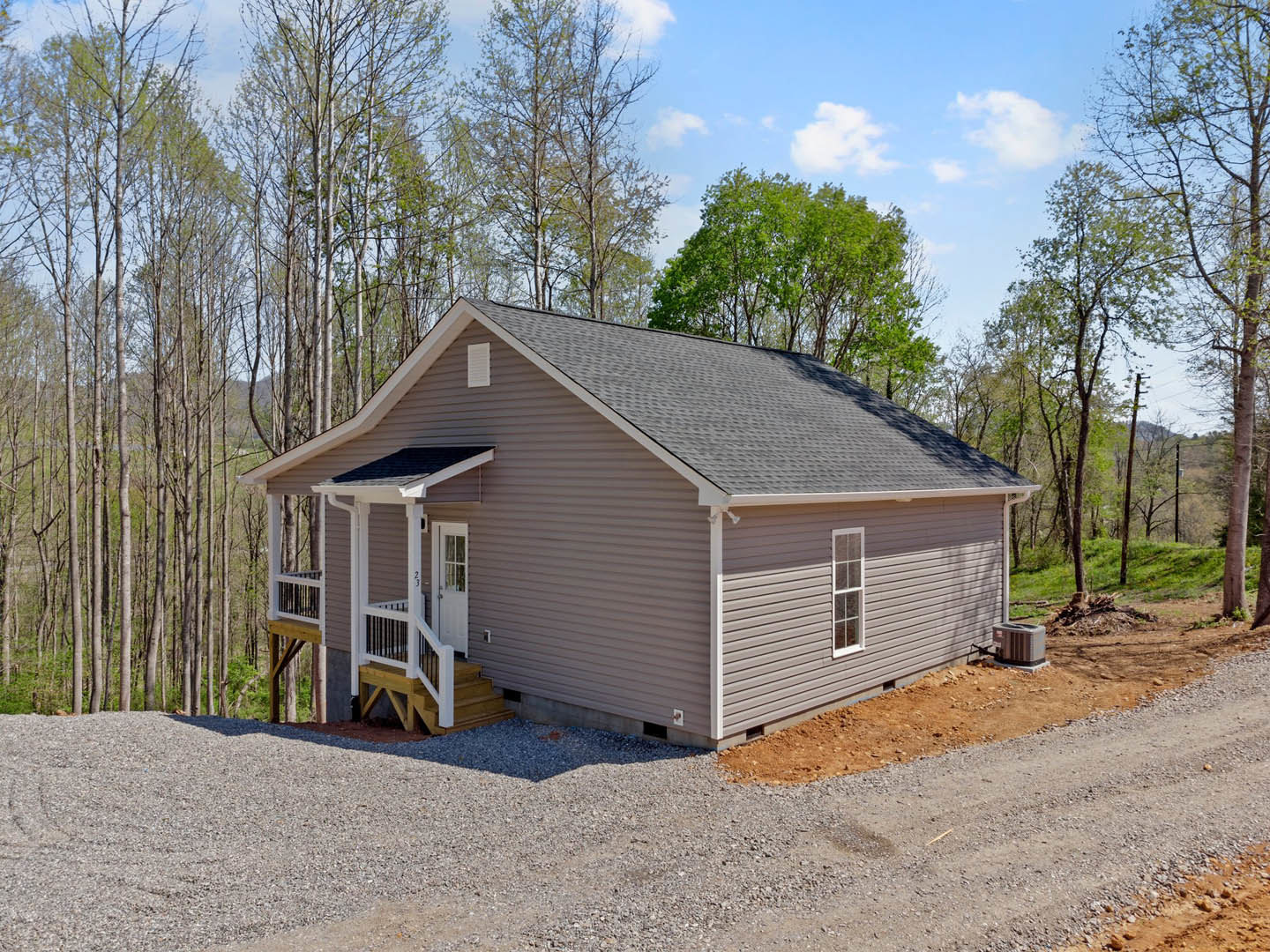 Two-story cottage with white porch railing, gravel driveway, and mature trees; grey heater unit sits beside foundation, dirt landscaping surrounds the house.