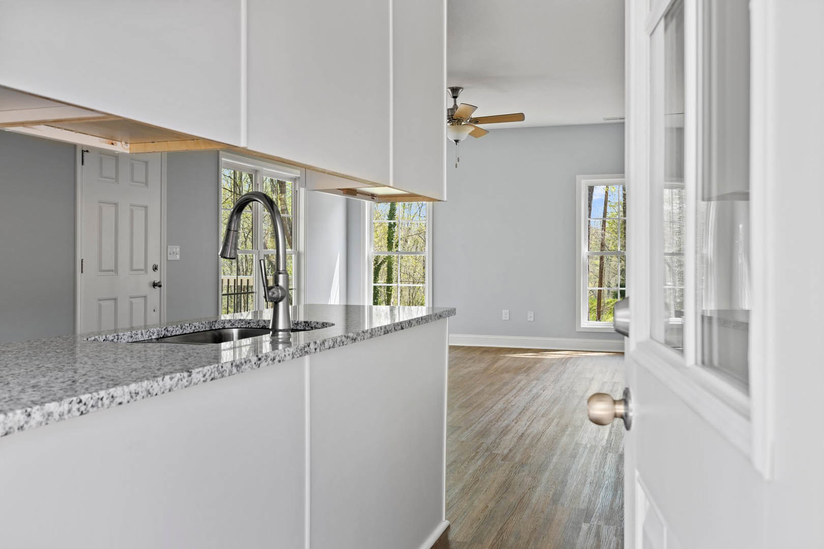 Modern kitchen with stainless steel sink, chrome faucet, wood plank flooring, white cabinetry, stone countertop, and large window overlooking leafy tree
