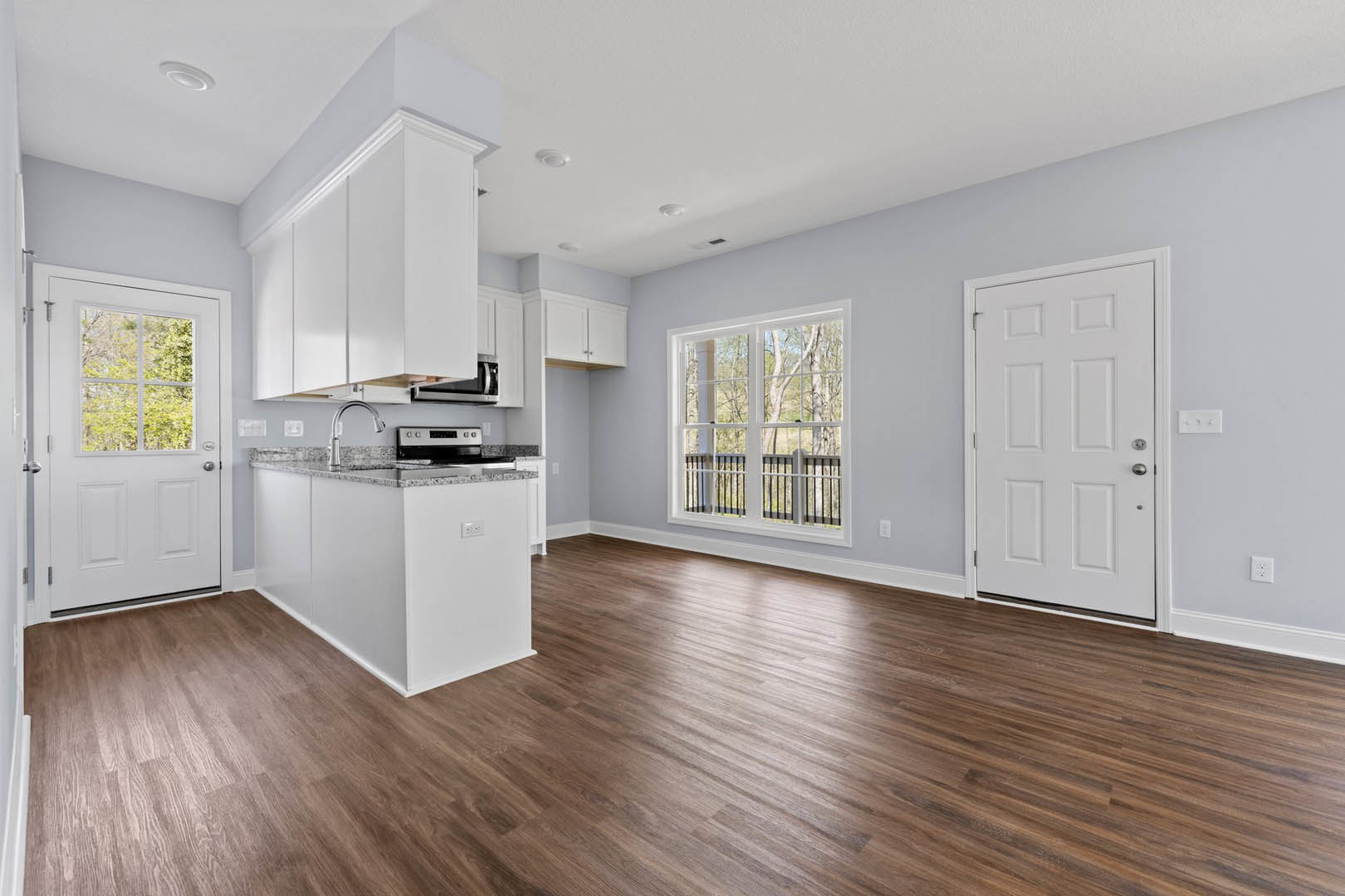 Open kitchen and dining area with hardwood floors, marble countertop, white cabinetry, stainless steel appliances, white door with silver handle, large window overlooking trees