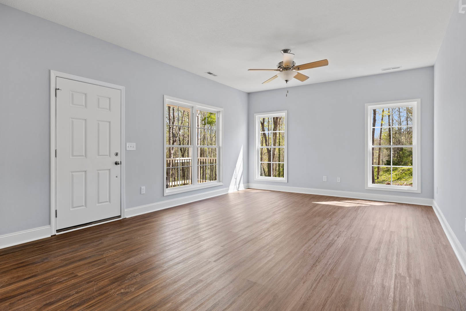 Wood flooring room with ceiling fan, white door with sign, large windows showing trees outside