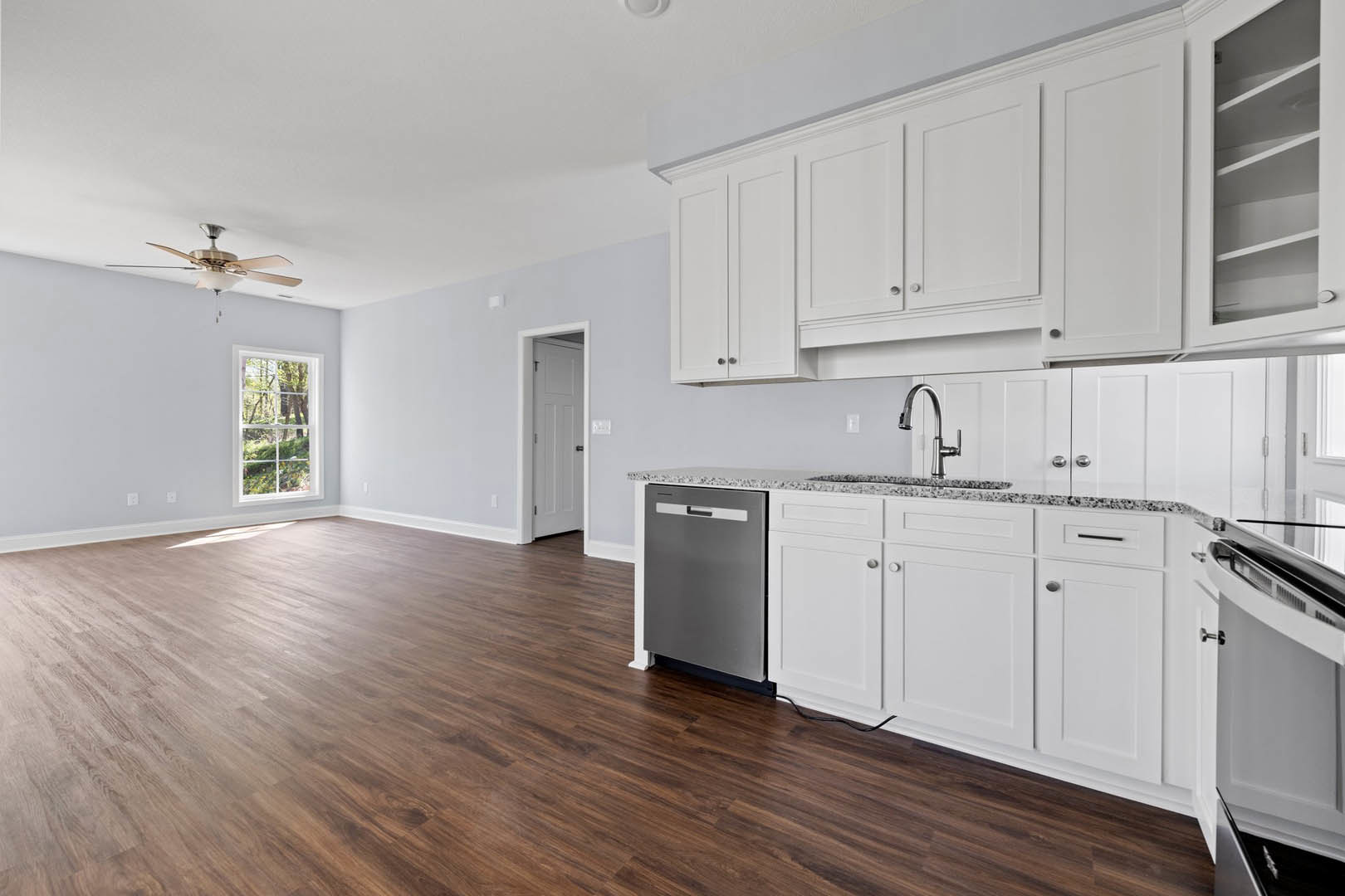 White kitchen cabinets, wood flooring, large window overlooking forest, ceiling fan with light fixture, white doors with silver and black hardware, grey rectangular appliance on