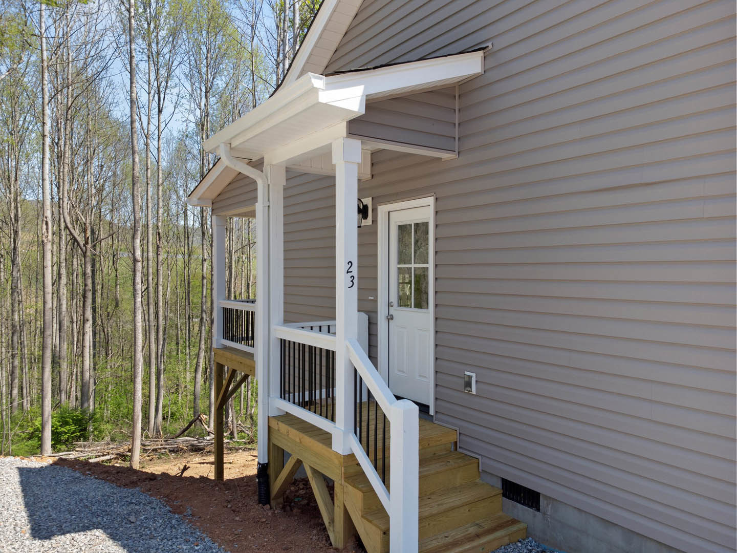 Front porch with white door, wooden stairs, light siding, gravel and dirt landscaping, trees in background, adjacent fence