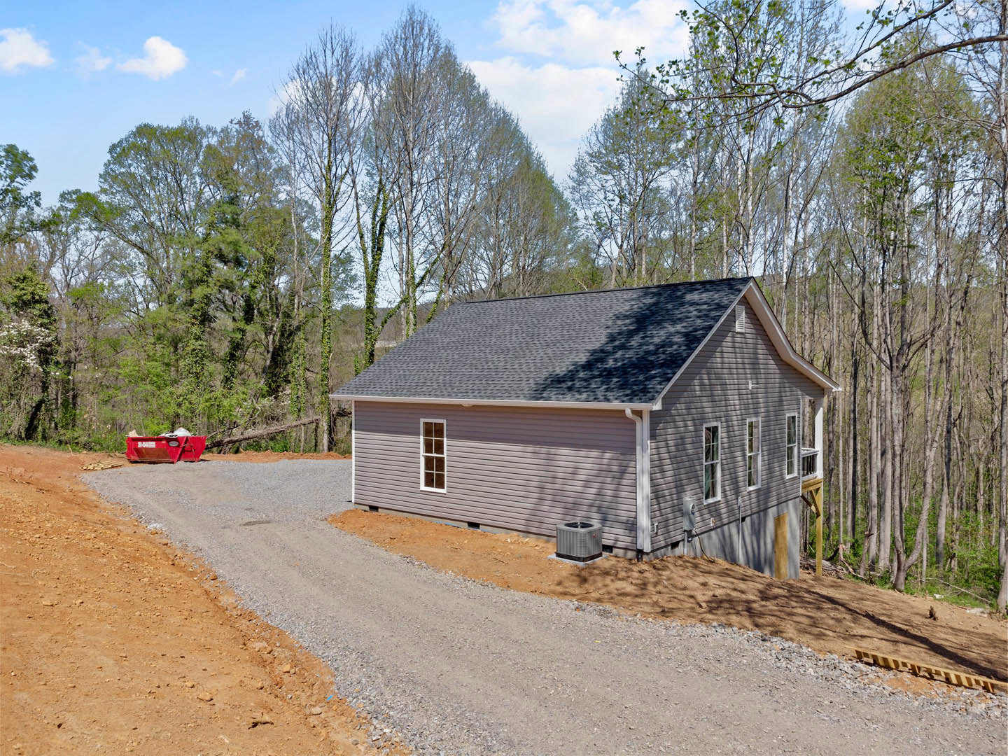 Two-story house with grey roof and white siding, gravel driveway, red dumpster, surrounded by tall green trees under blue sky with scattered clouds