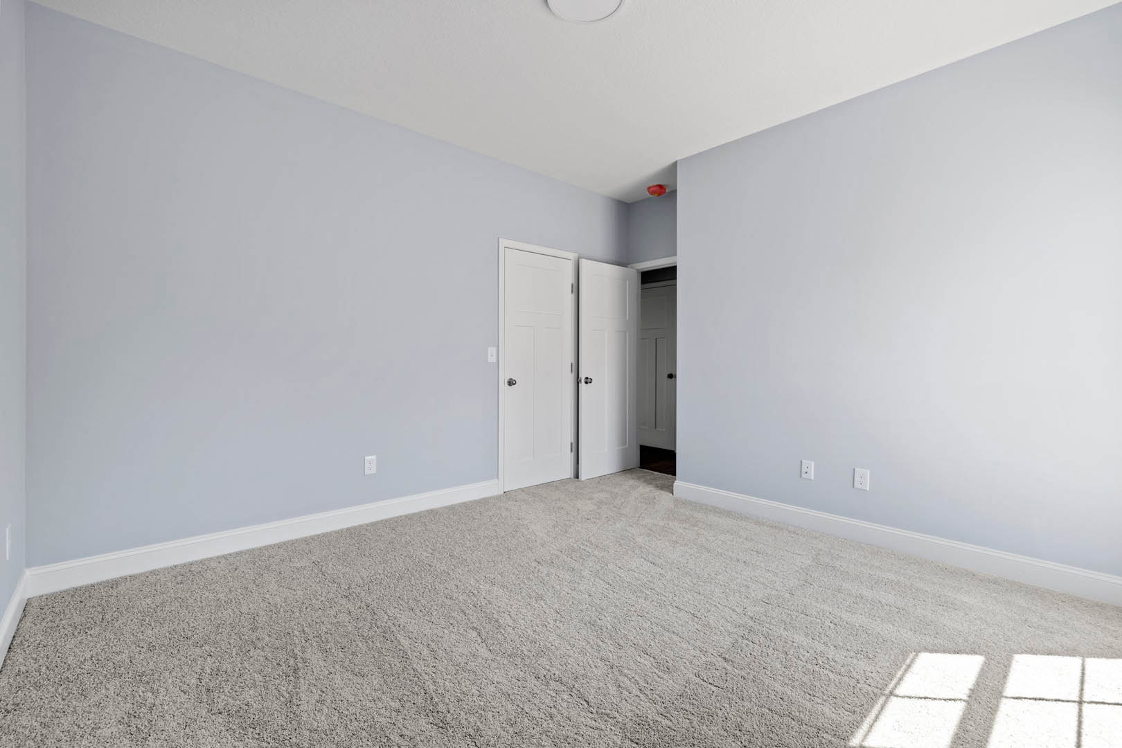 Carpeted room with white walls, open white door featuring a silver or black knob, sunlight casting window shadow on wall