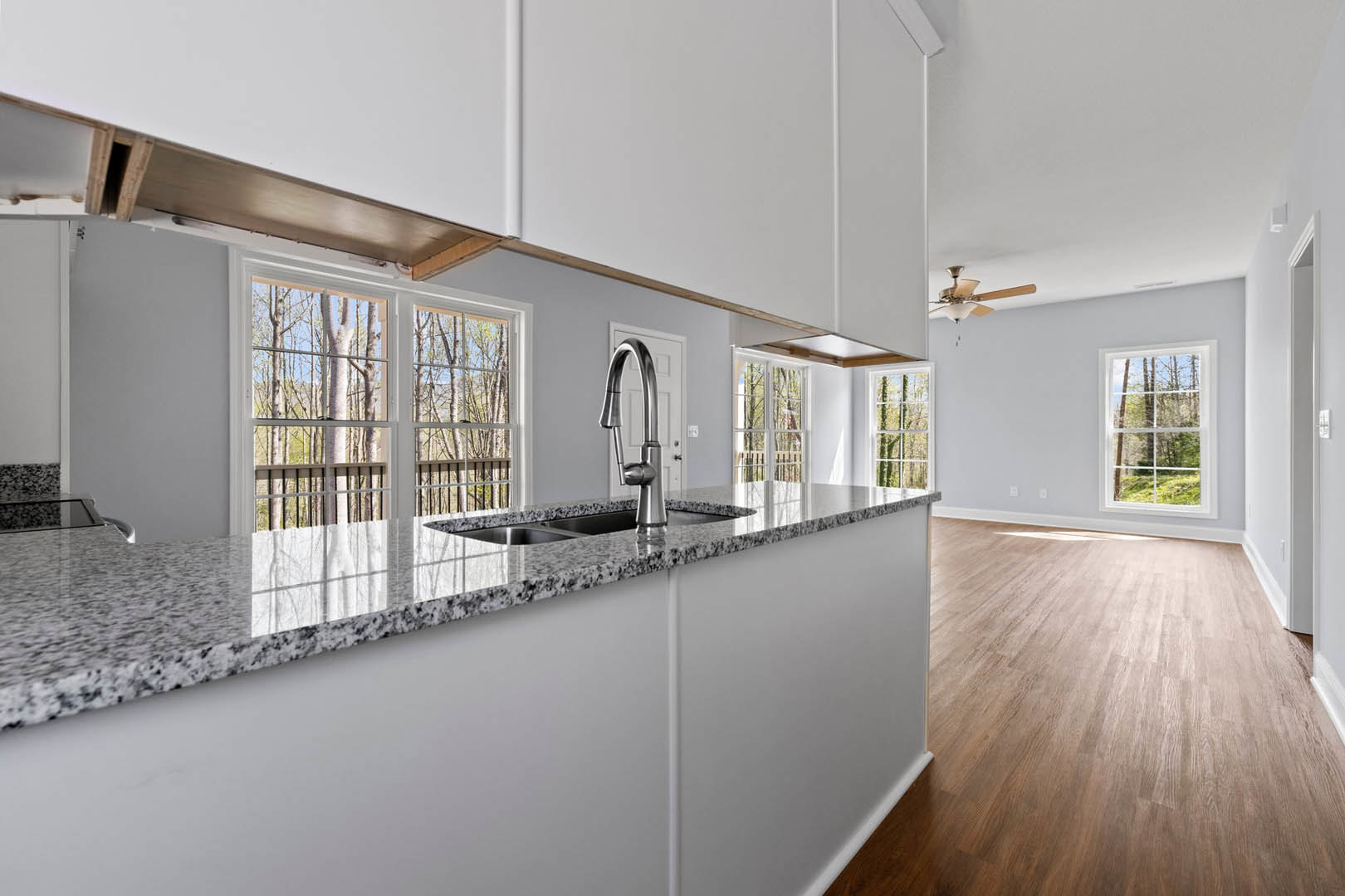 Kitchen with wood flooring, white walls, large window overlooking forest, stainless steel sink and faucet set in light-colored countertop, modern cabinetry.
