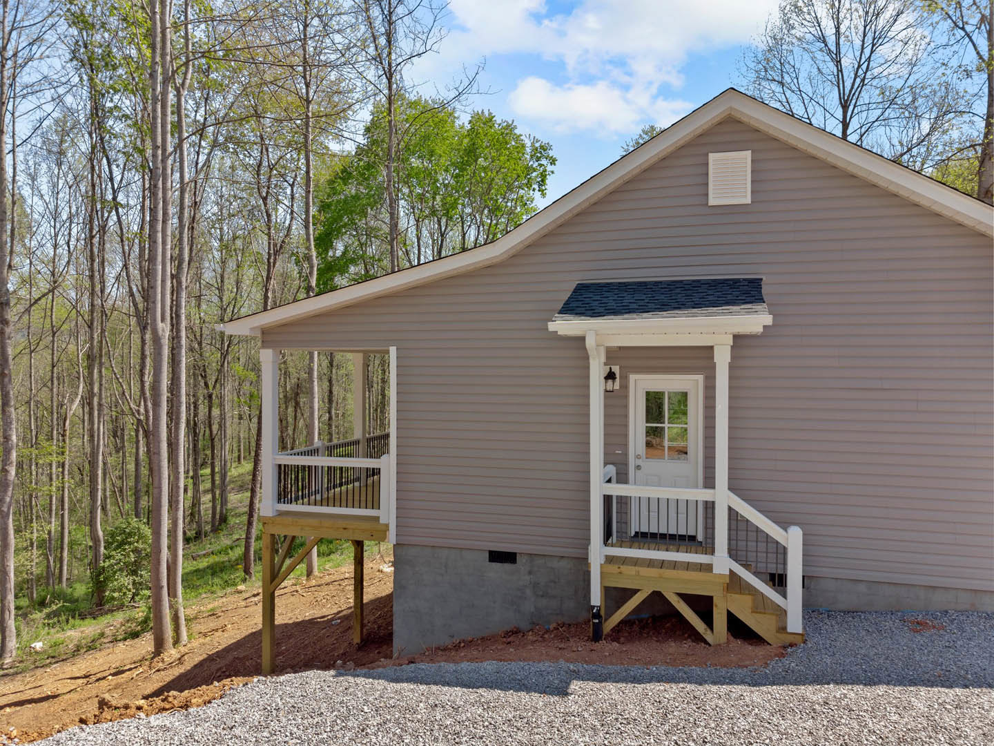 Two-story house with light siding, elevated wooden porch, white railing, glass-paneled front door, and stairs leading up the hill, surrounded by trees and cloudy sky.
