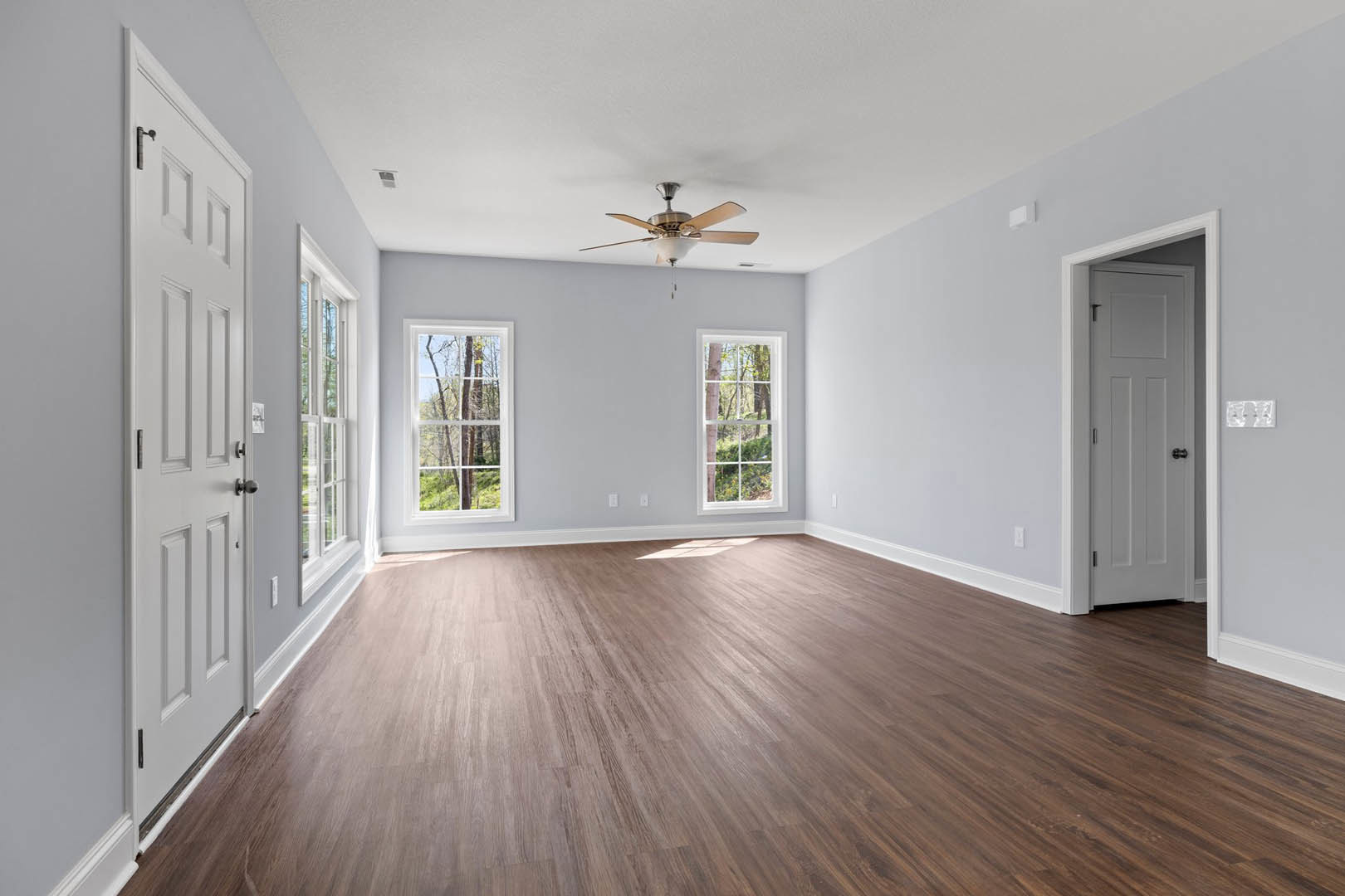 Hardwood floor room with ceiling fan and light fixture, large windows showing trees outside, white door with black handle, neutral walls and trim