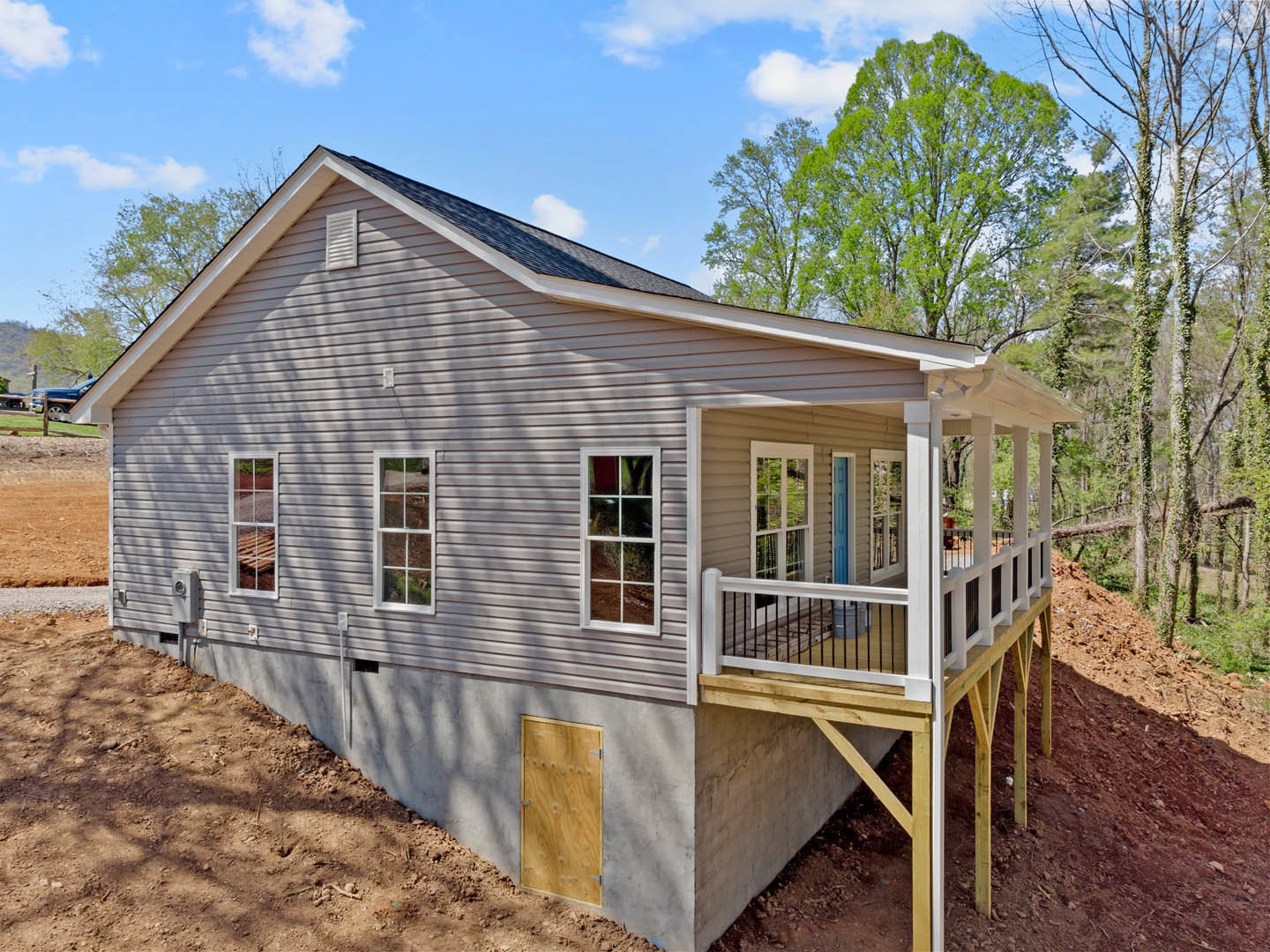 White house with covered porch and wooden deck, situated on a grassy hill with trees and cloudy sky in background; features white-framed windows and a wooden front door.