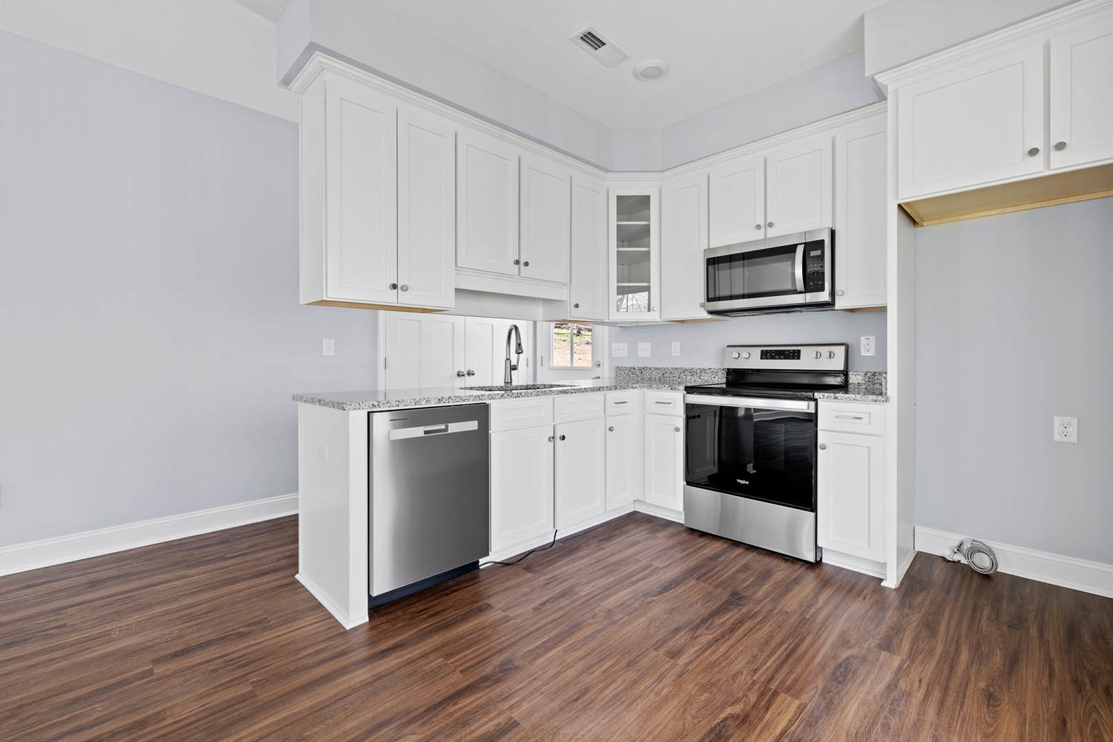 Kitchen with white shaker cabinets, stainless steel appliances including stove, microwave, refrigerator, and dishwasher, wood plank flooring, black cord visible on floor, light