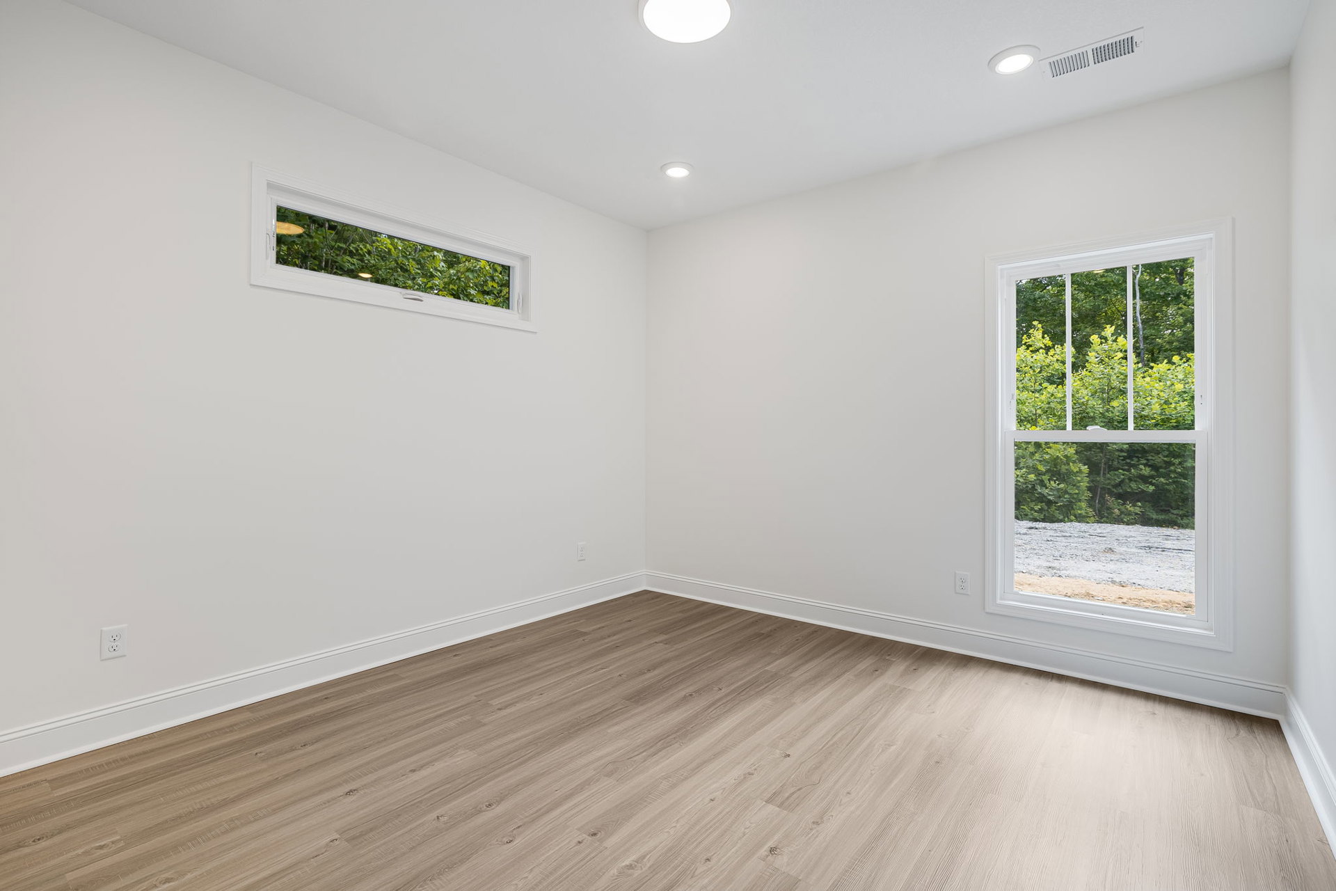Hardwood floor room with white plaster walls, large window framing leafy trees, wall vent visible near baseboard