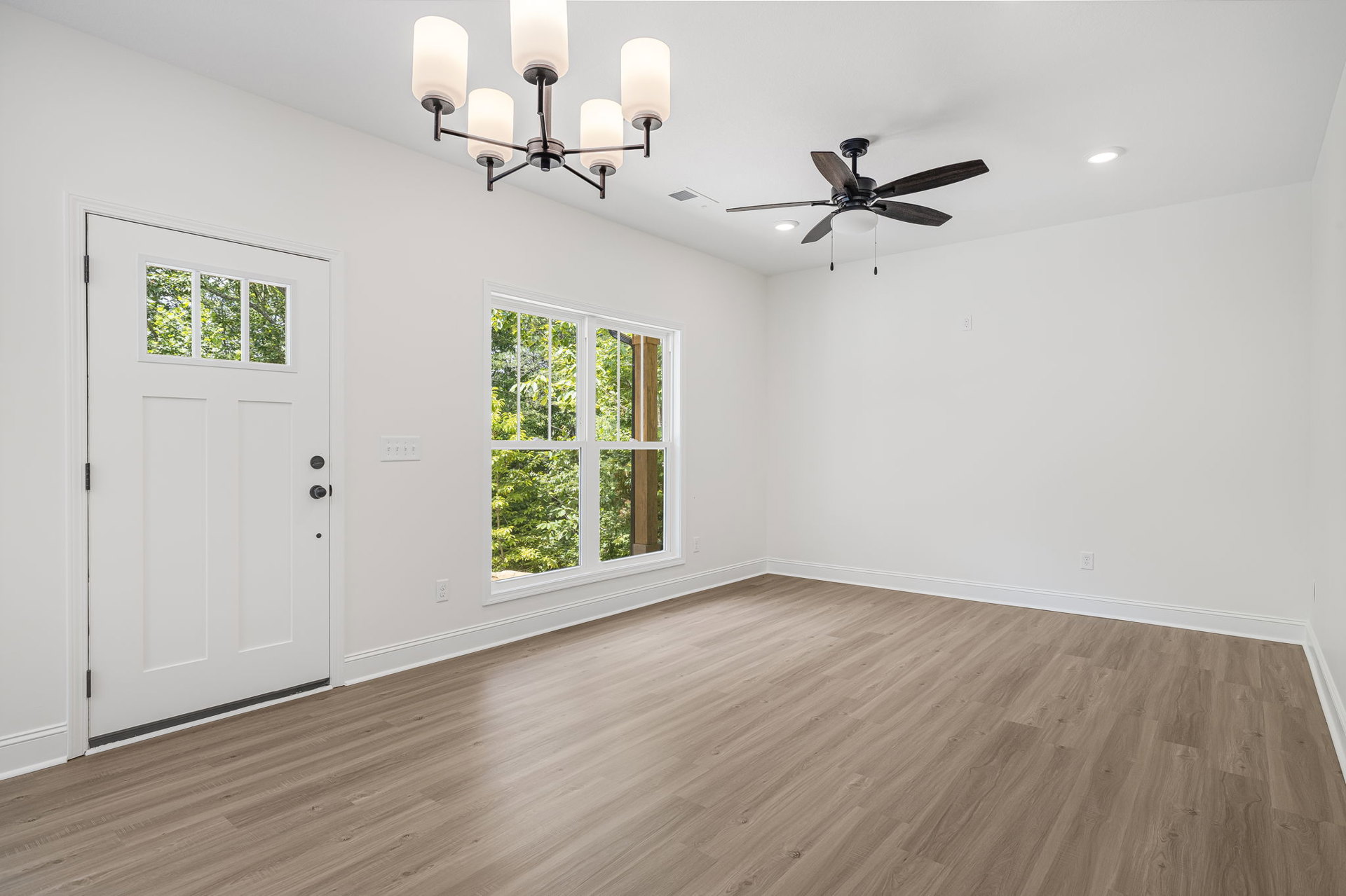 Spacious room with wood flooring, white walls, ceiling fan with light, large windows showing trees outside, and a white door with glass panel