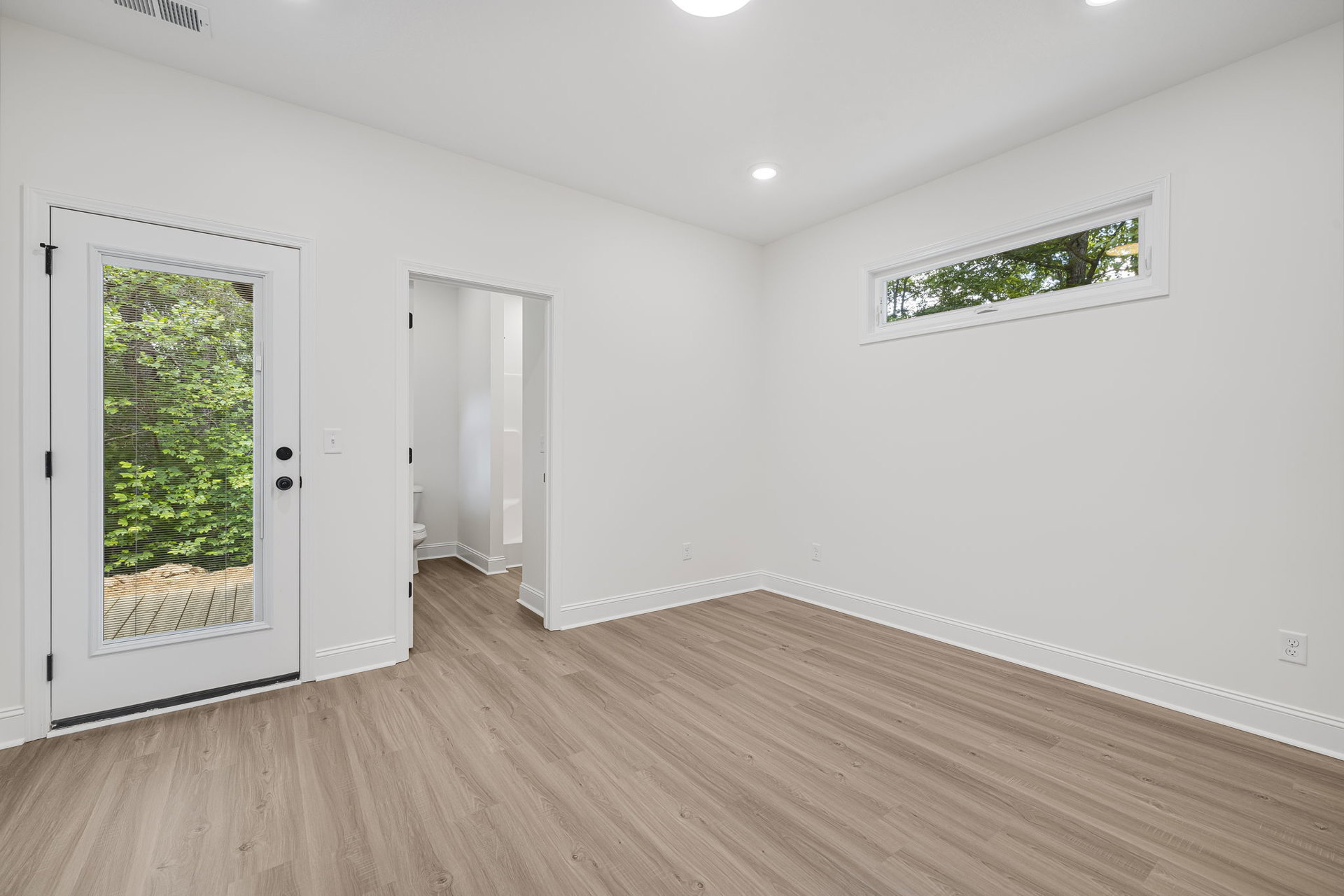 Wood flooring and white plaster walls in a room with a door and large window, green tree and rocks visible outside