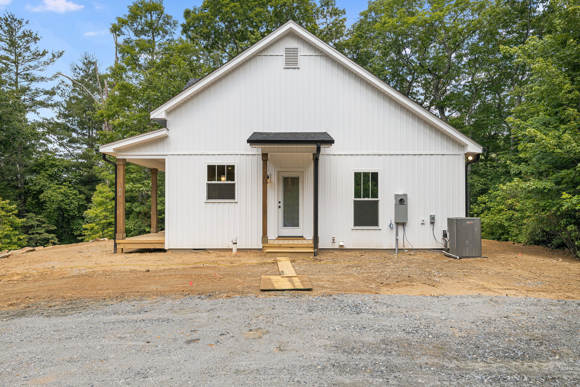 White siding house with black awning over front door, gravel driveway, wood planks on ground, single window illuminated, surrounded by mature trees.