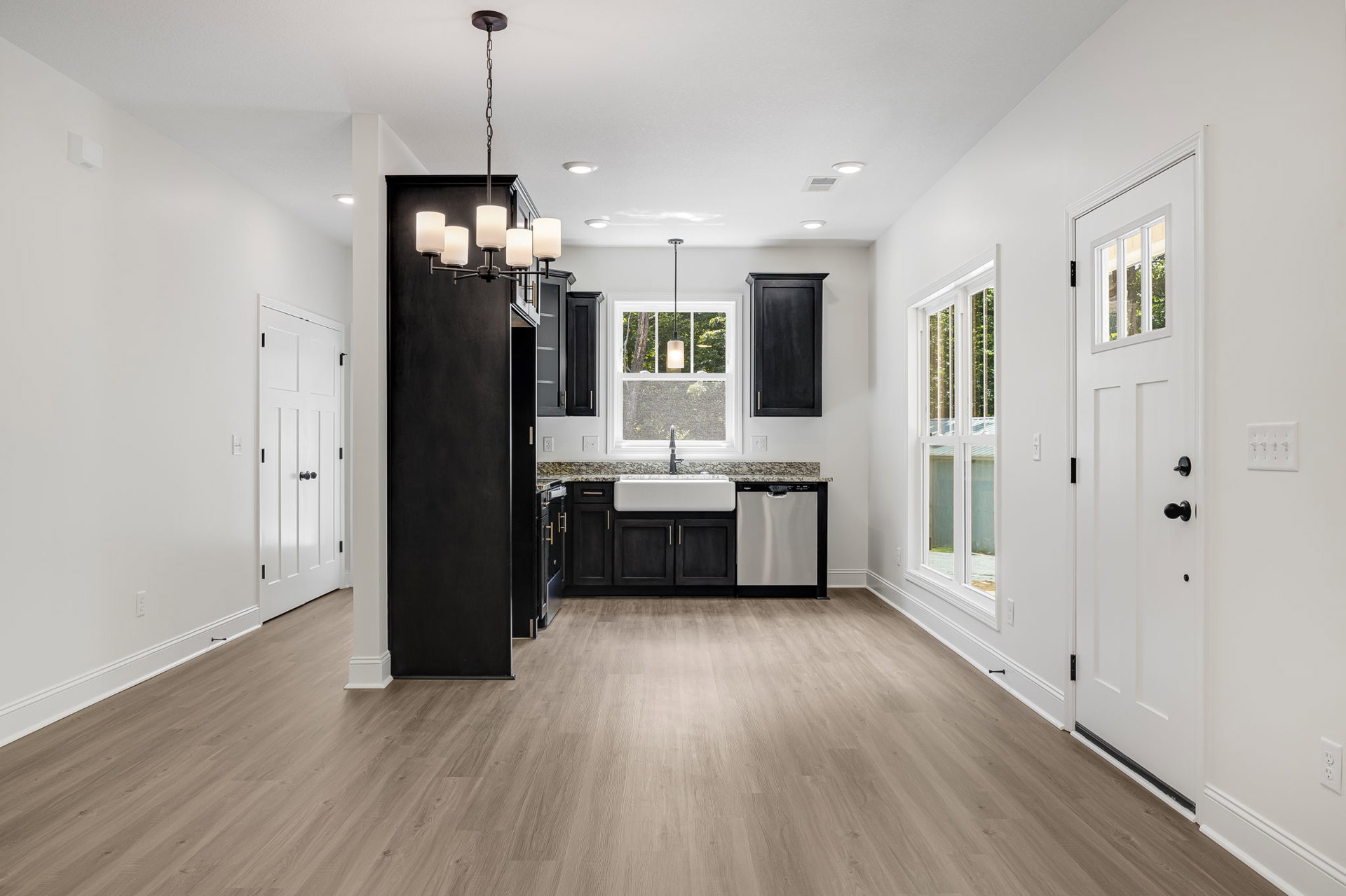 Hardwood floor kitchen with white cabinetry, central sink, ornate chandelier, black door with white trim, white door with black knobs, and window letting in natural light