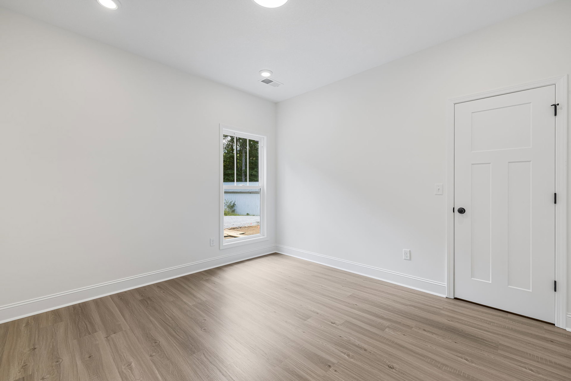 Hardwood floor room with white door featuring black knobs, white-framed window showing trees outside, ceiling vent, and recessed light.