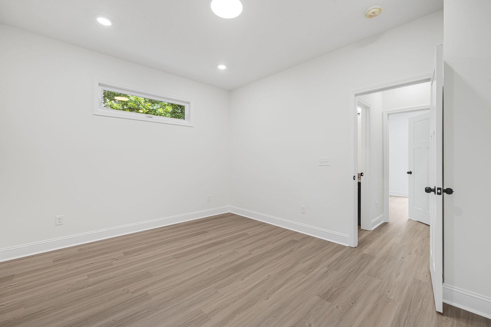 Room with hardwood floor, large window showing trees, white ceiling with recessed lights, white door featuring black handles, smooth plaster walls