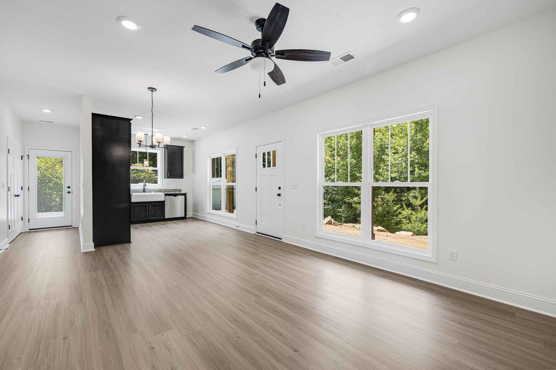Ceiling fan with light fixture, wood laminate flooring, white door with window, black wall-mounted board with light, large window showing trees outside