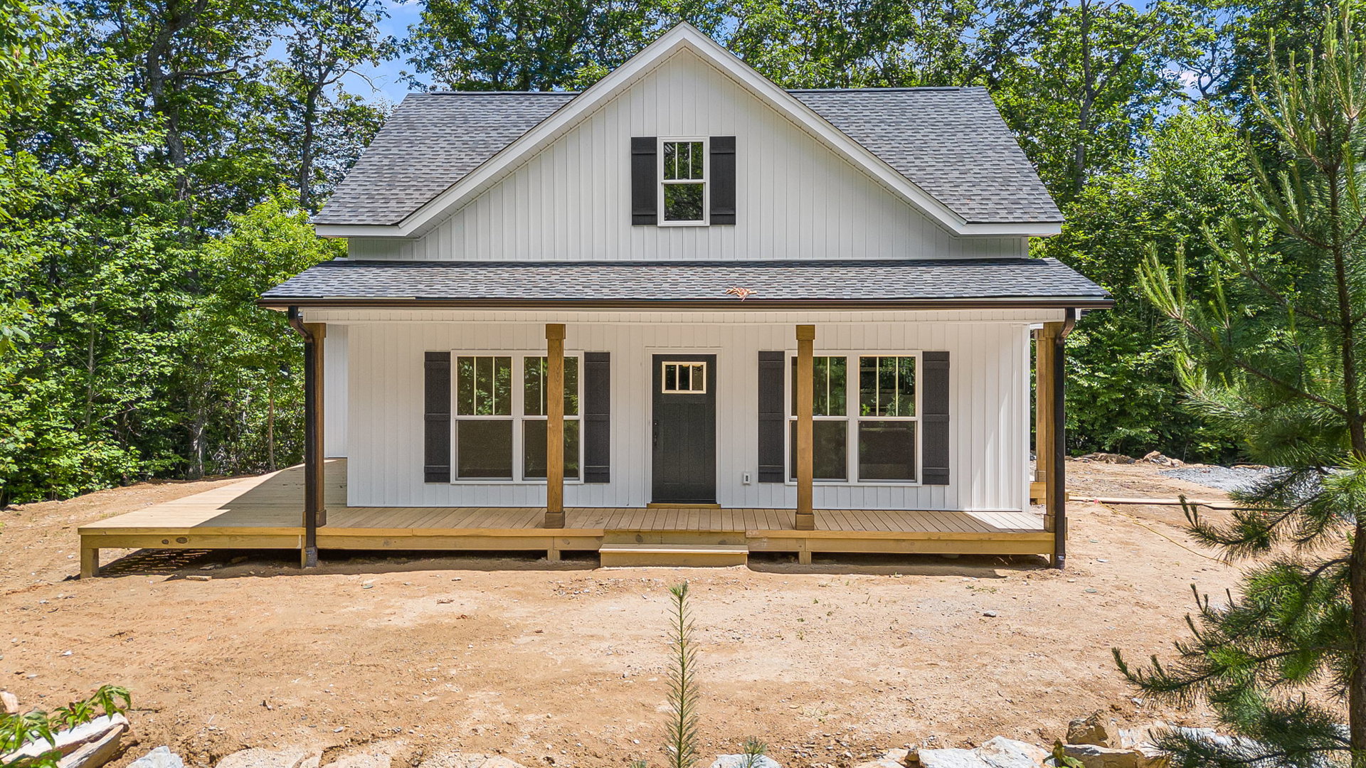 White siding house with black front door, window with dark shutters, covered porch, small tree planted in dirt, simple landscaping
