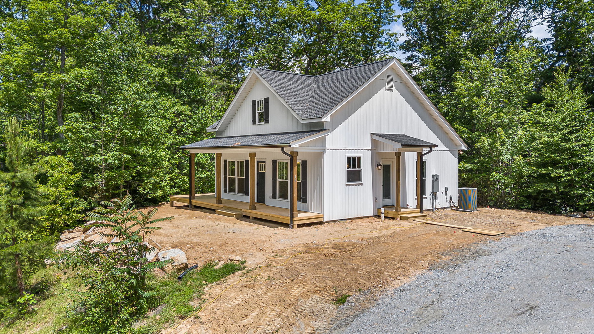 White siding house with covered porch, gravel driveway, green lawn, mature trees in background, black pipe in grass, blue and yellow striped utility box, close-up window with white