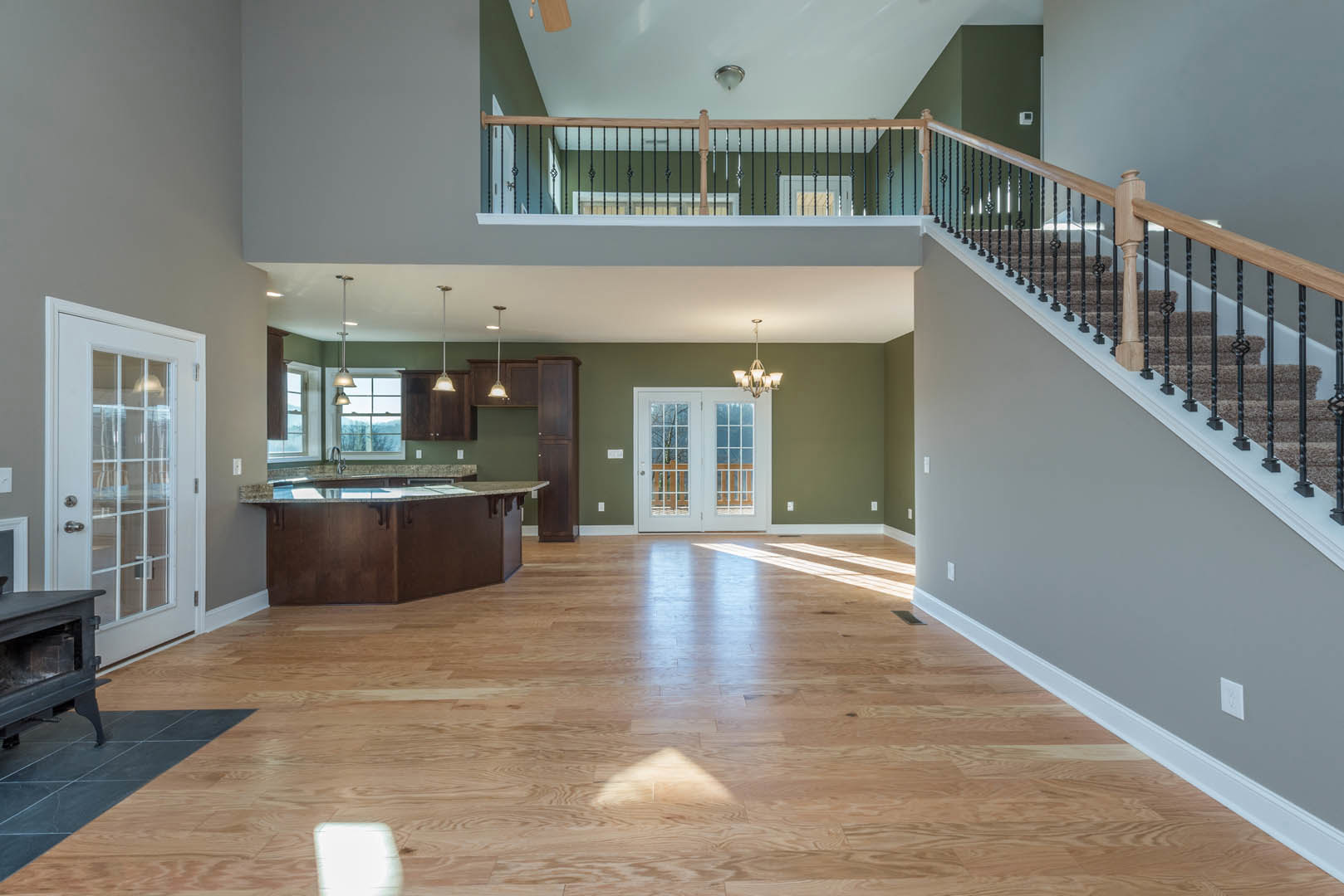 Open-concept room featuring hardwood floors, a modern kitchen with white cabinetry, a staircase with wooden railing, double glass-paneled doors, and a wood stove near a white door