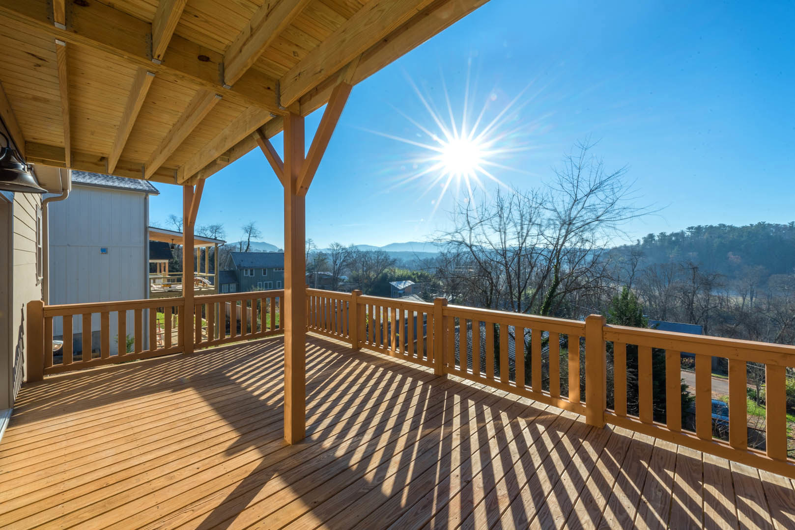 Wooden deck with railings overlooking mountain landscape, sunlight streaming through clear blue sky, shadows cast across outdoor flooring, trees visible in background