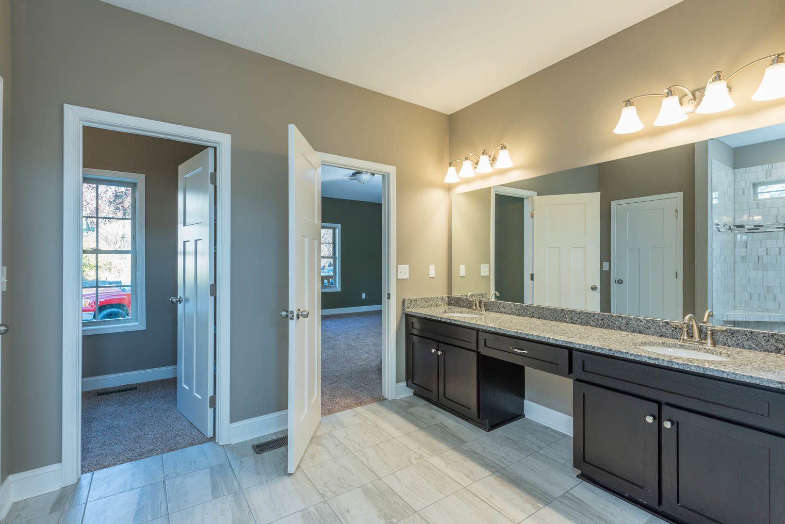 Double sink vanity with white countertop, large wall mirror, white door with silver knob, window overlooking trees, two-lamp light fixture above mirror, white tiled shower with
