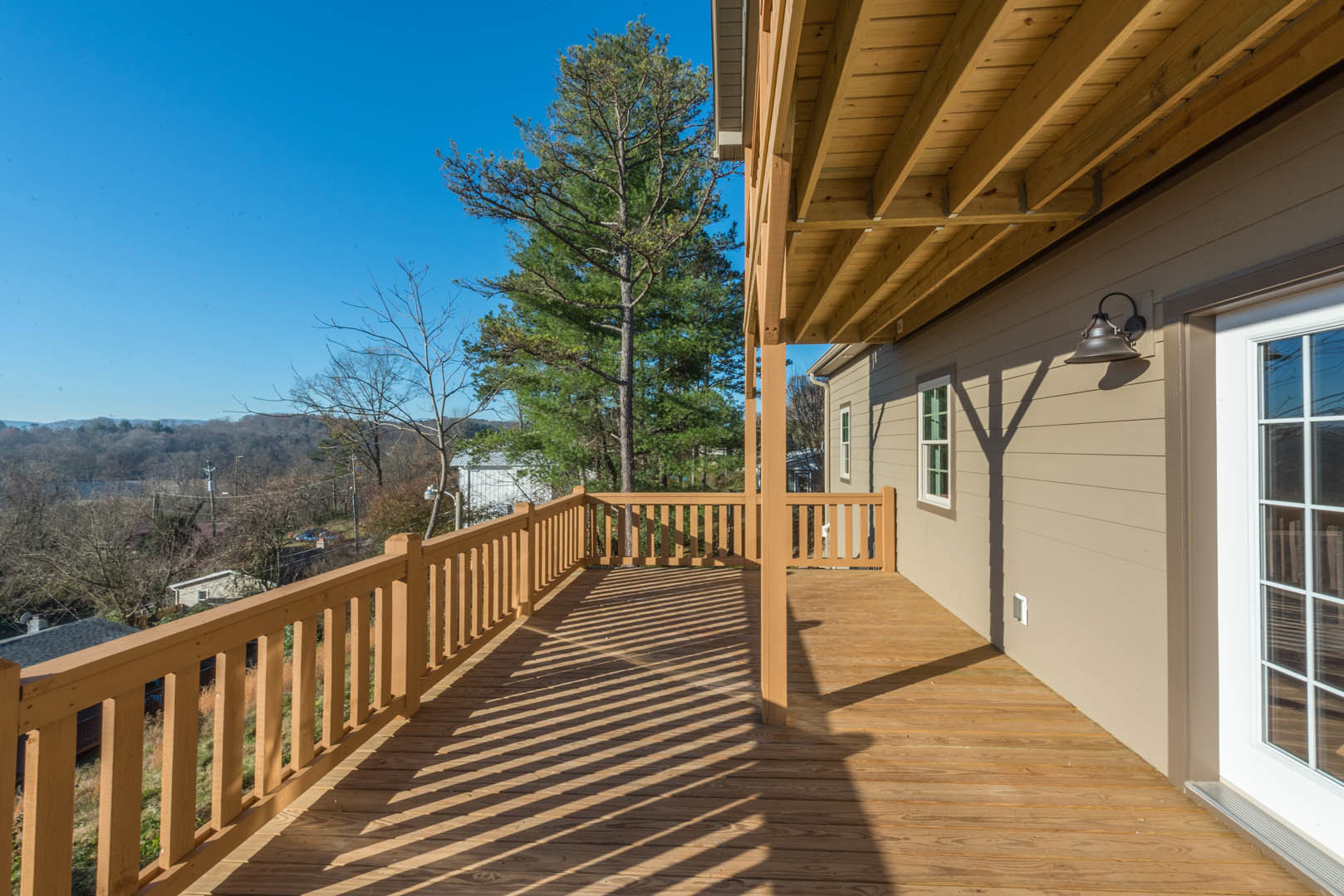 Wooden deck with railings and shadows, tall tree with green leaves, blue sky, close-up of window and outdoor light fixture