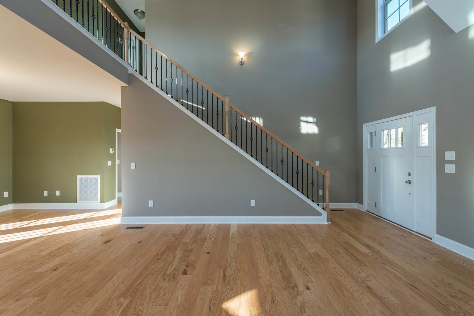 Wood staircase with black metal railing, light wood flooring, white walls, white door with silver handle, wall-mounted light fixture, and window with white trim.