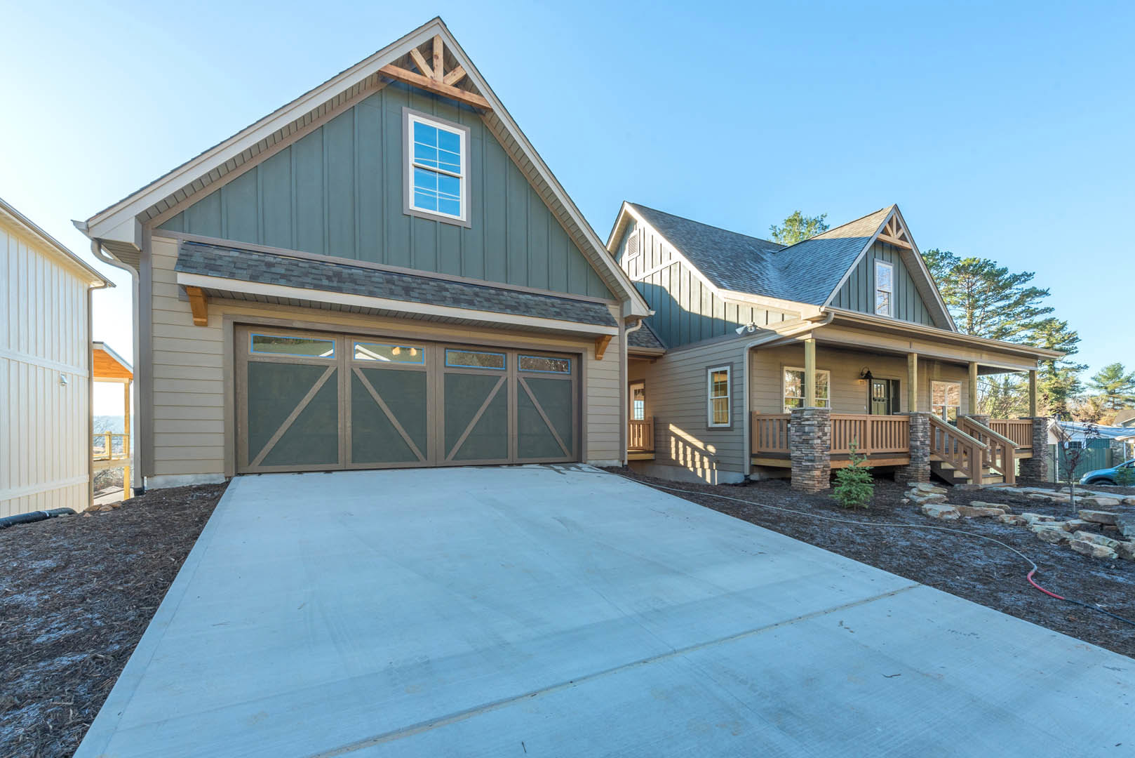 Two-story home with light siding, prominent garage door, paved driveway, large front window, small tree planted near entry, gabled roof against blue sky