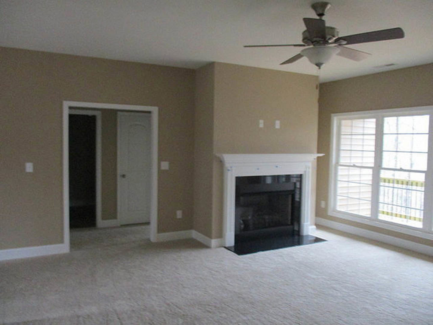 Living room with light-colored walls, stone fireplace, ceiling fan with light fixture, white-framed window, and white door.