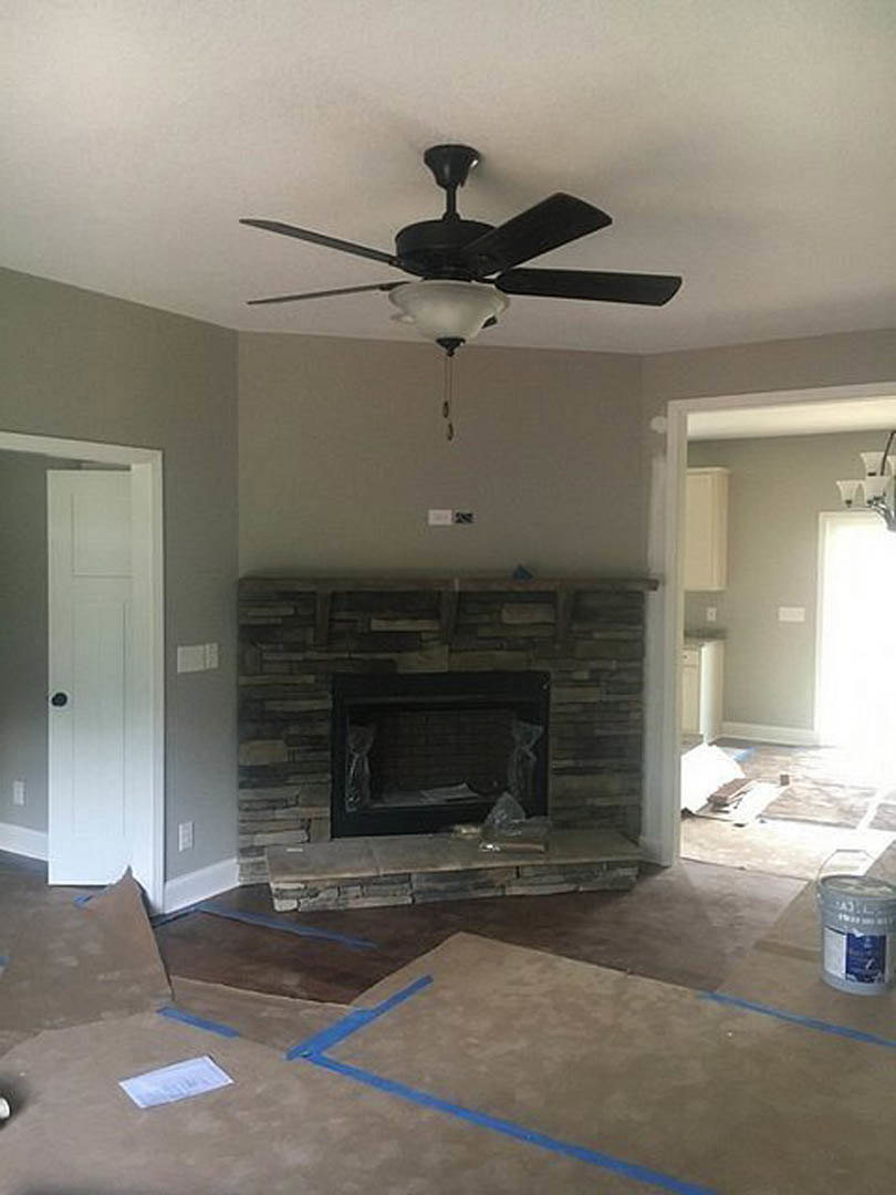 Living room with ceiling fan and light fixture, brick fireplace wrapped in plastic, white container on hardwood floor, plaster walls and ceiling.