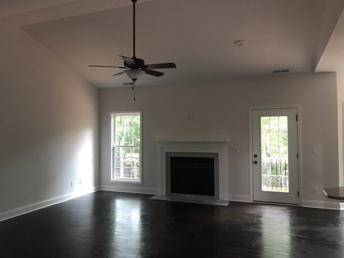 Living room with white plaster walls, dark hardwood flooring, stone fireplace, ceiling fan with light, large window showing trees, and glass-paneled door.