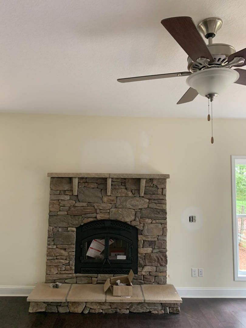Stone fireplace with glass door and black trim, ceiling fan with light fixture above, white door and railing on the side, hardwood floor, neutral walls.