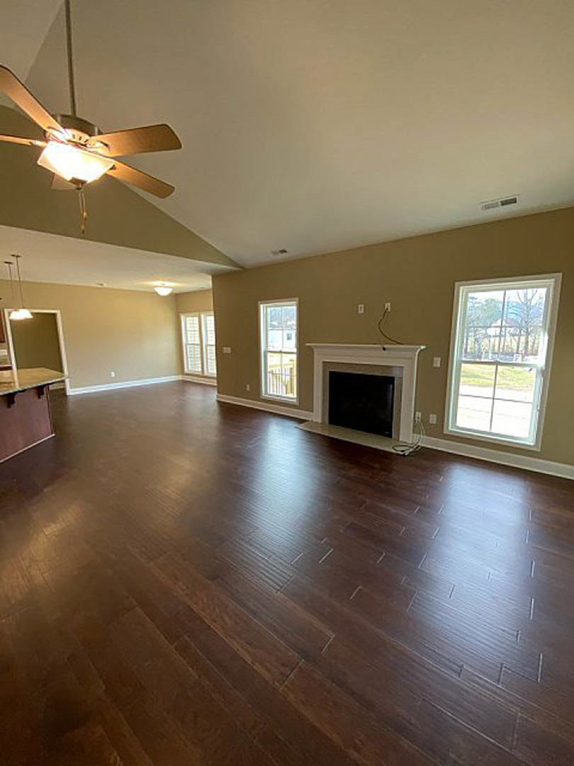 Living room with hardwood flooring, white fireplace mantle, ceiling fan with light, large window showing neighboring house, black square object on white surface