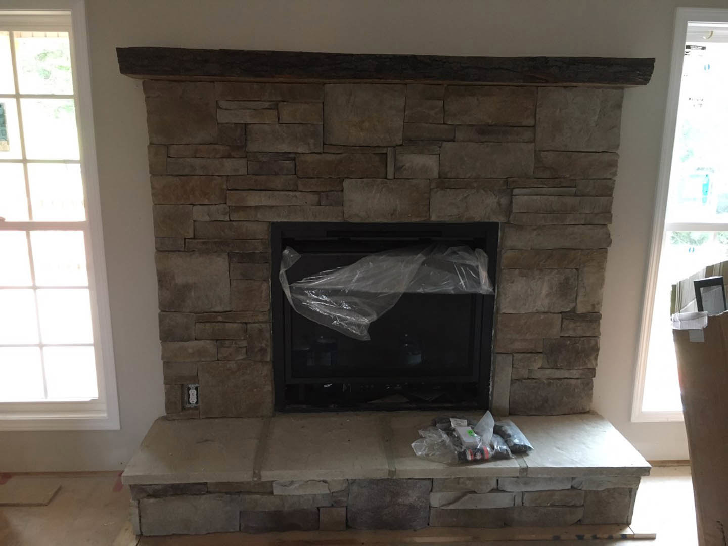 Stone fireplace partially covered with plastic wrap, wood mantel shelf above, adjacent window with white trim, neutral painted walls in a cozy living room.