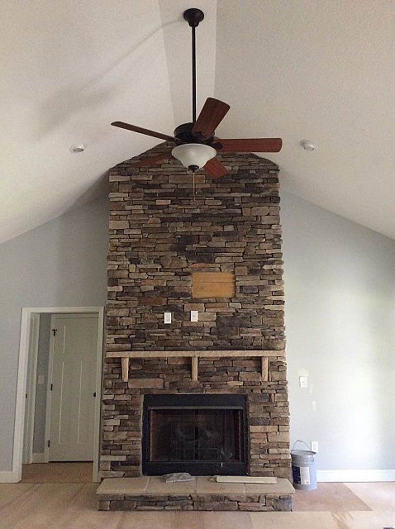 Ceiling fan mounted above a fireplace with a black frame and shelf, white door with black knobs, wooden beam on the wall, and brick wall visible in the background.