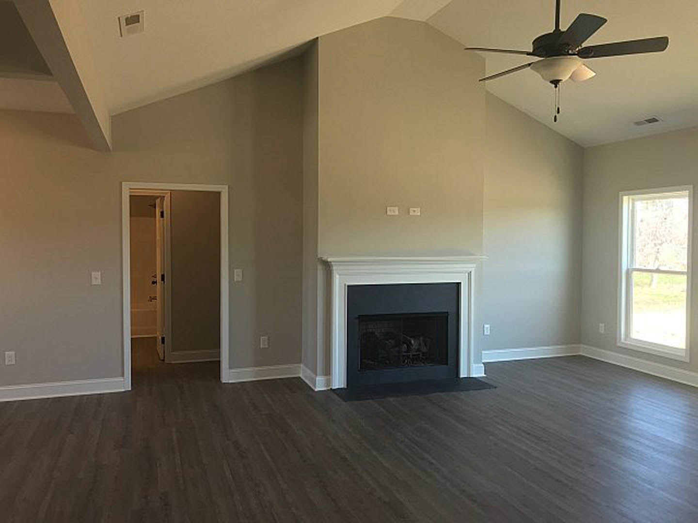 Living room with hardwood flooring, ceiling fan with light, glass-door fireplace, white-framed window, and white wall vent