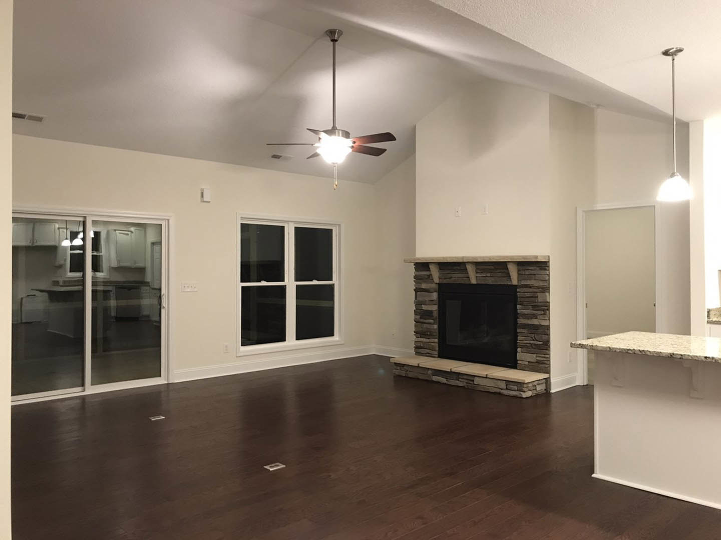 Living room featuring a black fireplace with glass window and stone ledge, ceiling fan with light fixture, sliding glass door with white frame, plaster walls, and wood flooring