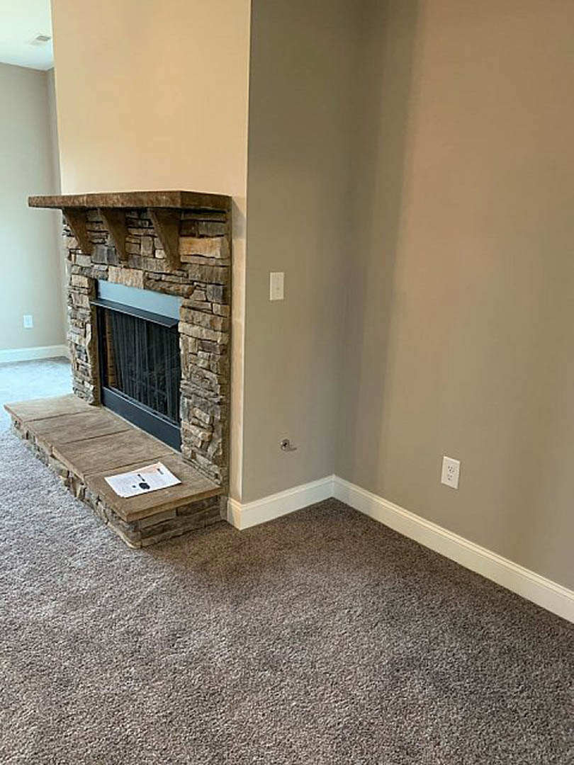 Stone fireplace with black fire screen set against textured wall, carpeted floor, window with white trim, and paper with black logo resting on a step