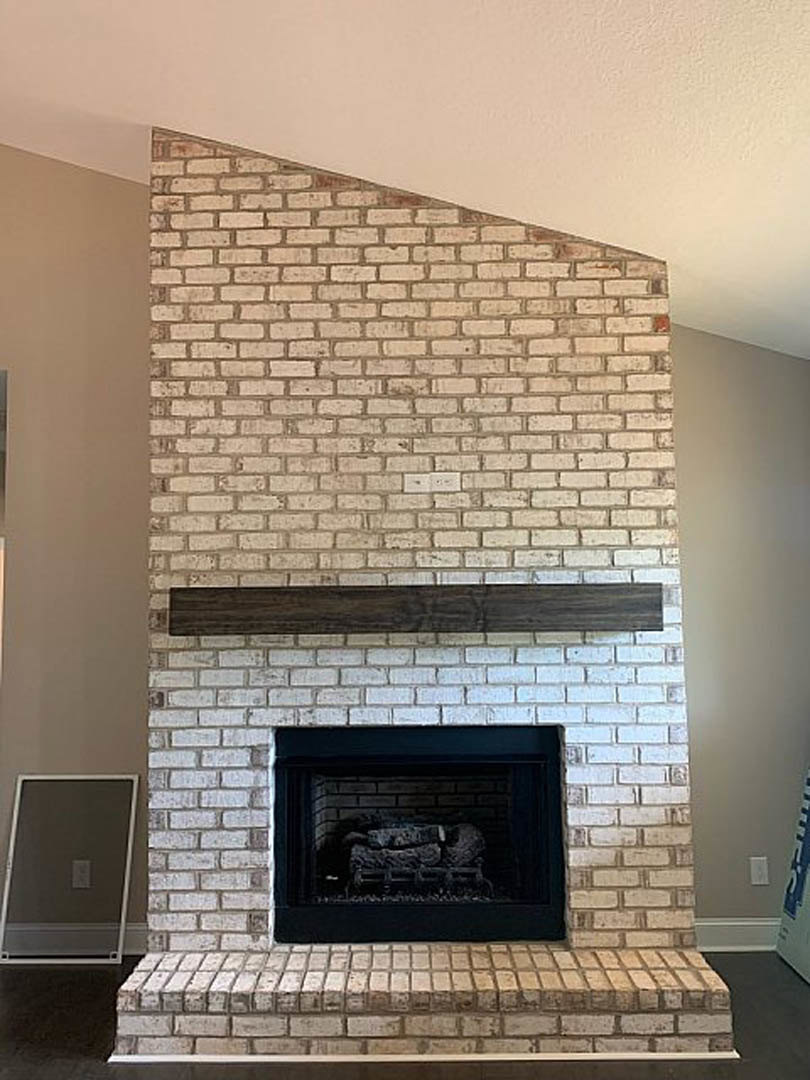 Brick fireplace with wood mantel, stacked logs in hearth, white walls, large mirror above mantel, wood flooring