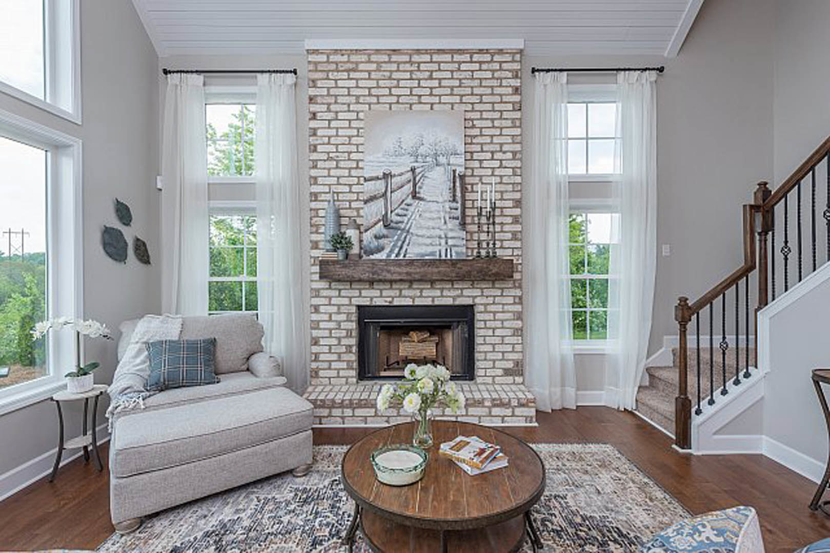 Living room with light-colored couch and matching footstool, stone fireplace, wooden coffee table holding a bowl, books, and white orchid in a pot, neutral walls and large window.